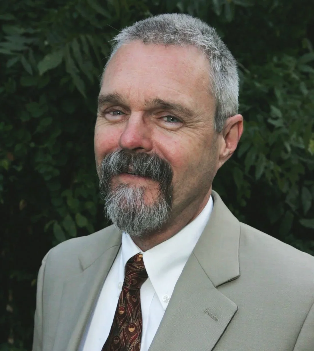 A middle-aged man with gray hair, a beard, and a mustache dressed in a beige suit, white shirt, and patterned tie, standing outdoors with green foliage in the background.