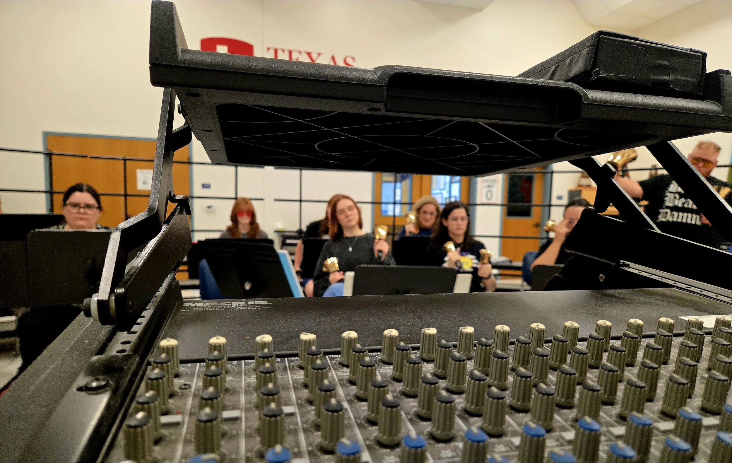 A music rehearsal space with a focus on a sound mixing board in the foreground. Out of focus in the background, a group of people are sitting in a semi-circle holding small golden bells, possibly practicing for a band or orchestra.