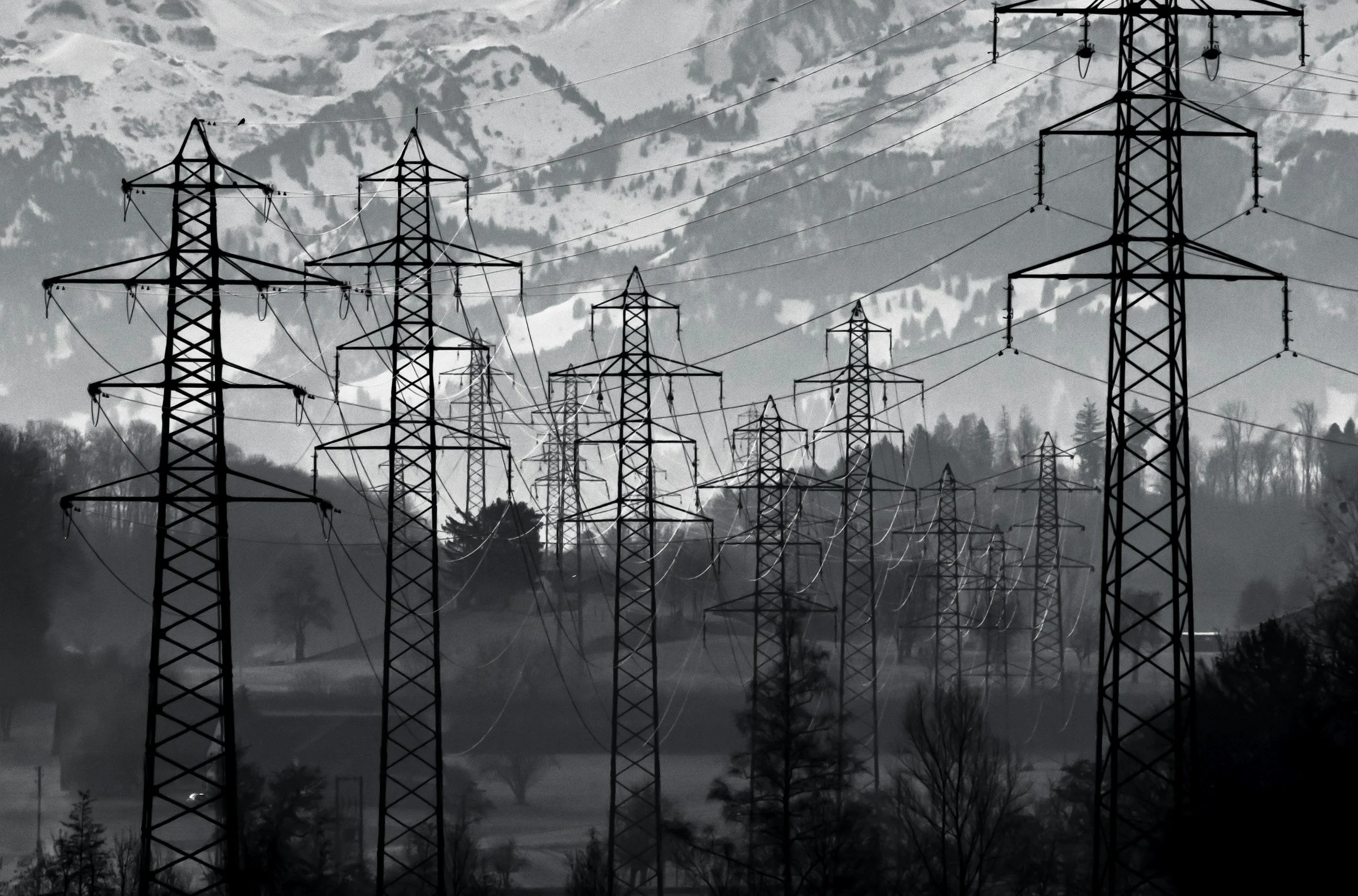 A series of electrical transmission towers in a mountainous landscape with snow-covered peaks in the background.