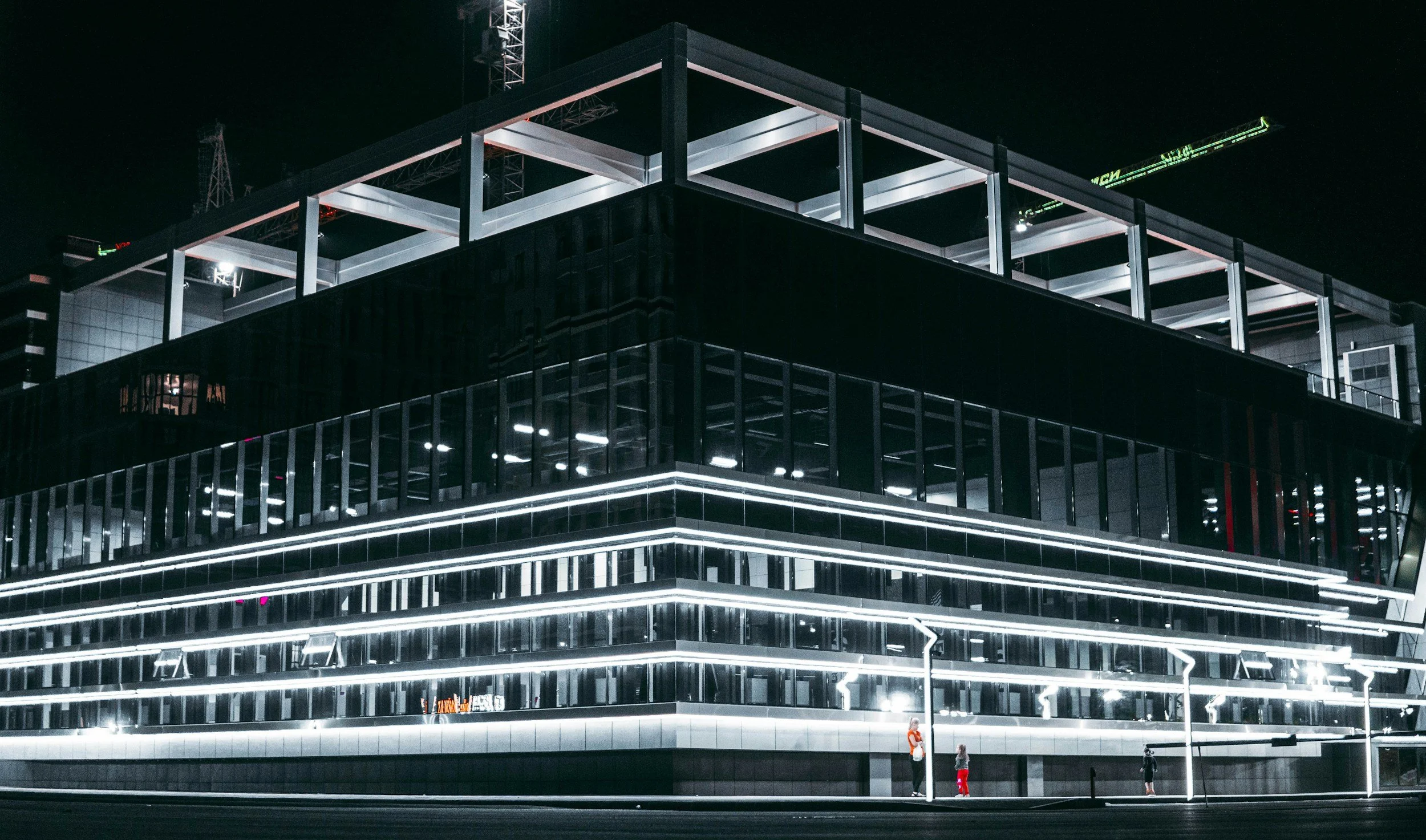 Night view of a modern building with illuminated horizontal lines on its facade. Three people are standing outside the building.