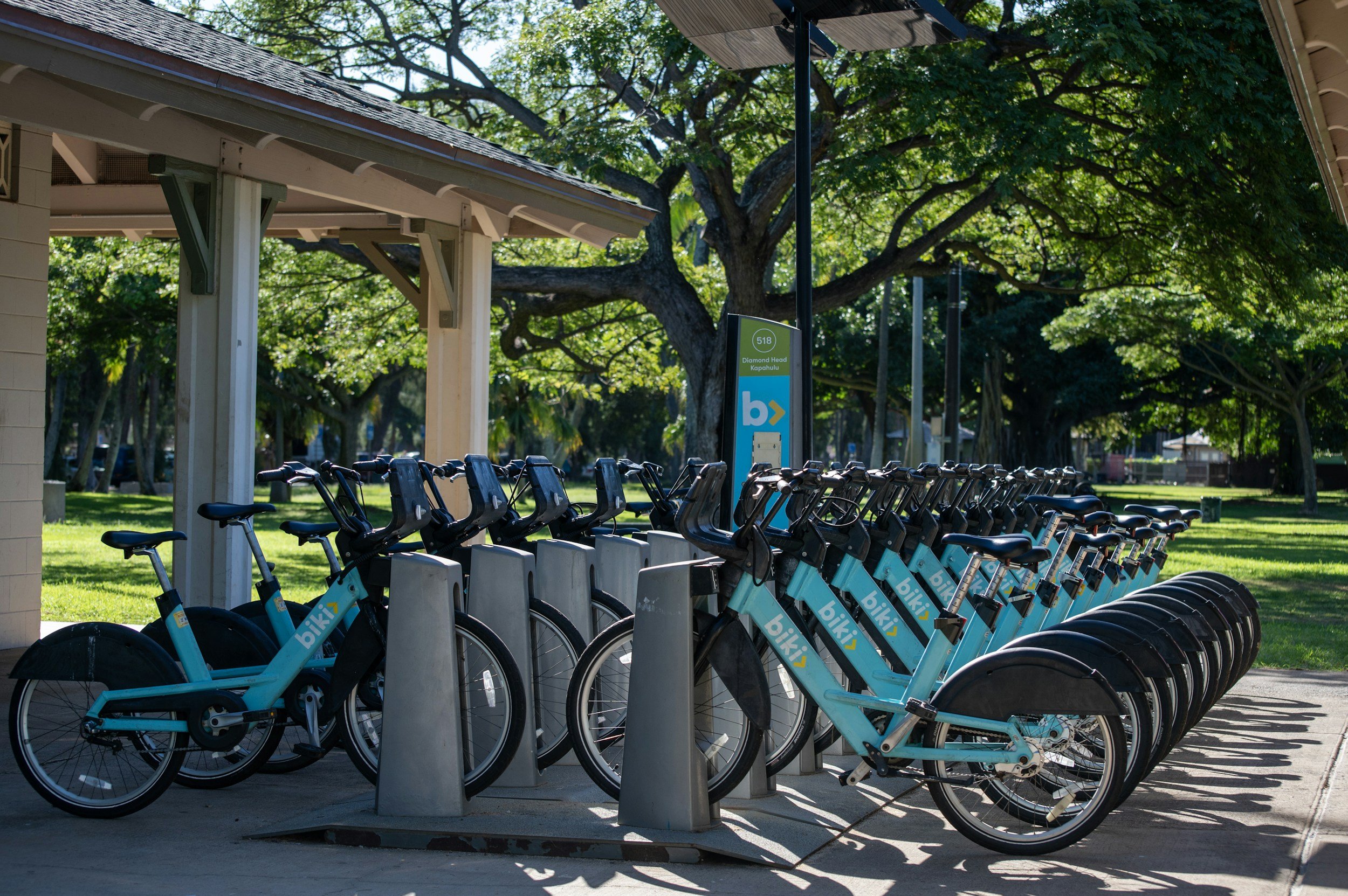 Row of blue Bik bike-sharing bicycles docked at a station in a park with green trees and a sign displaying the location as Diamond Head, Kapiolani.