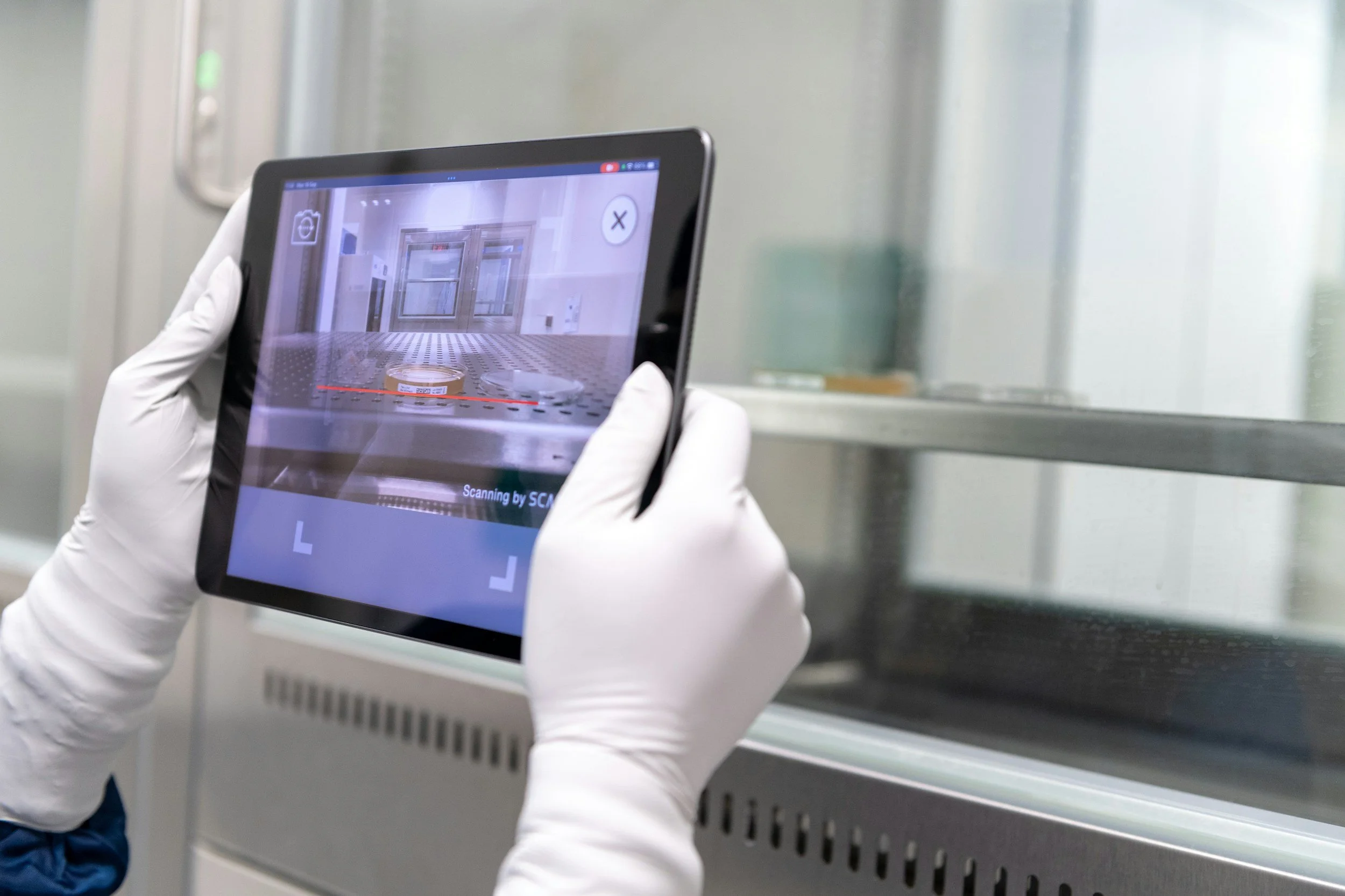 Person in white gloves scanning an object with a tablet in a laboratory or cleanroom environment.