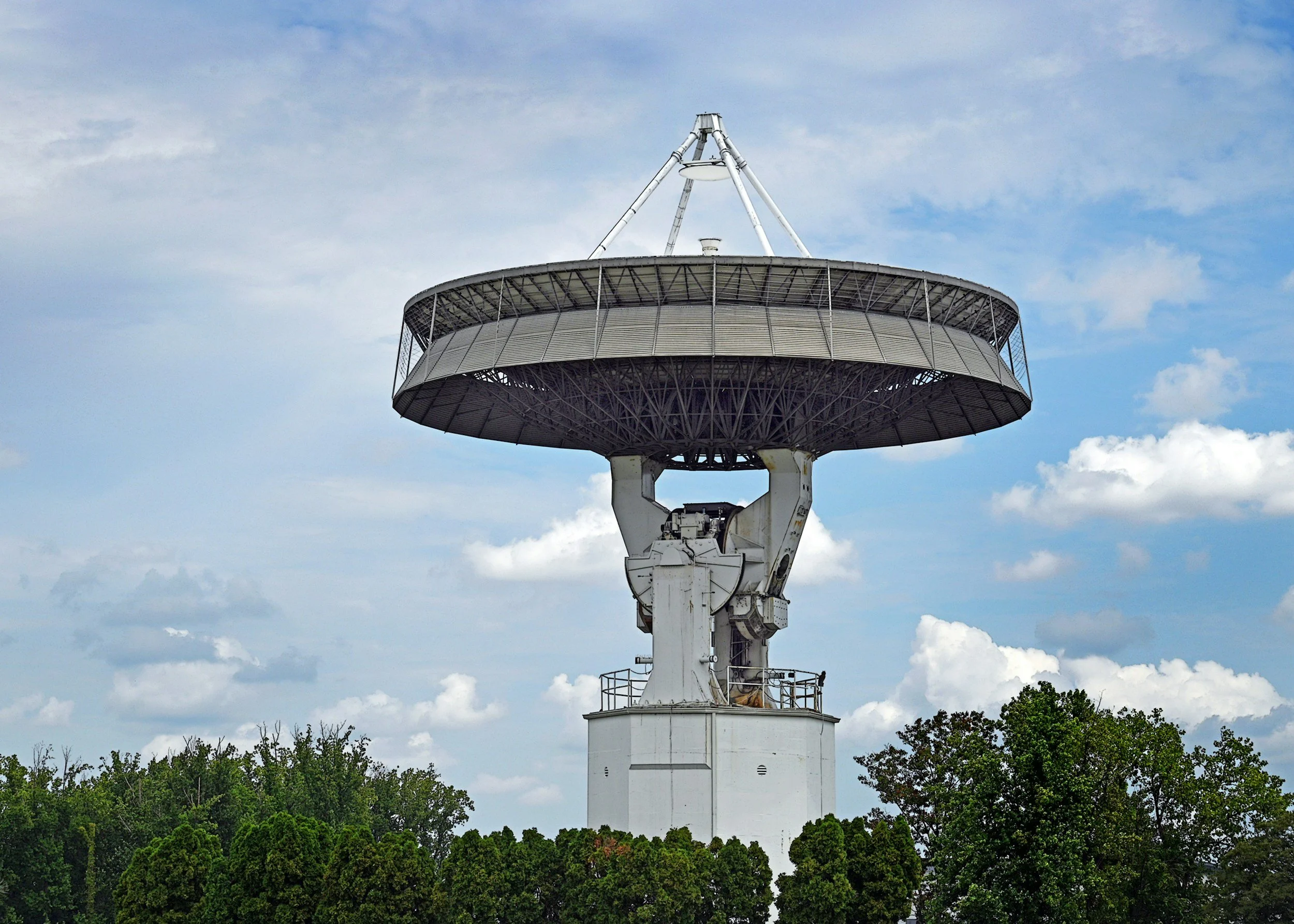 Large radio telescope with a circular dish and supporting structure, situated above trees under a cloudy sky.