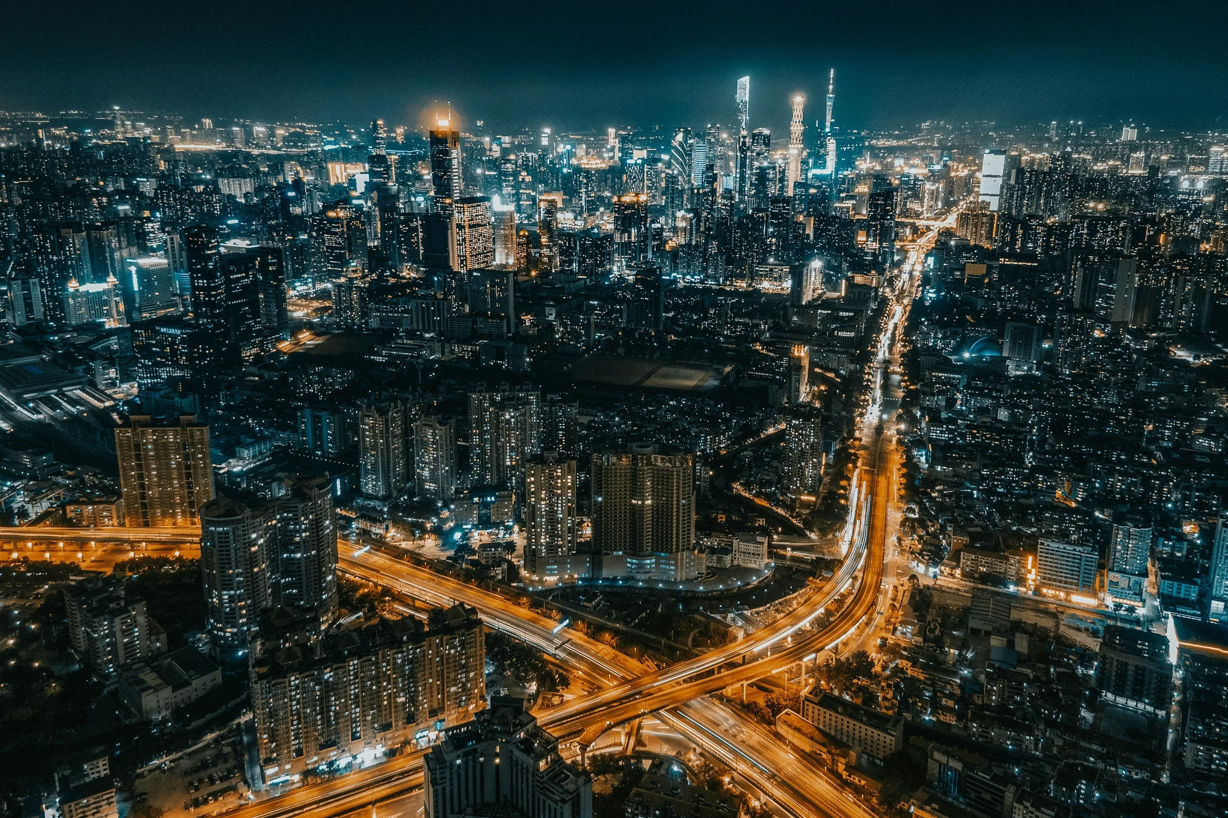 Nighttime aerial view of a city with illuminated skyscrapers and highways, with bright lights and tall buildings in the distance.