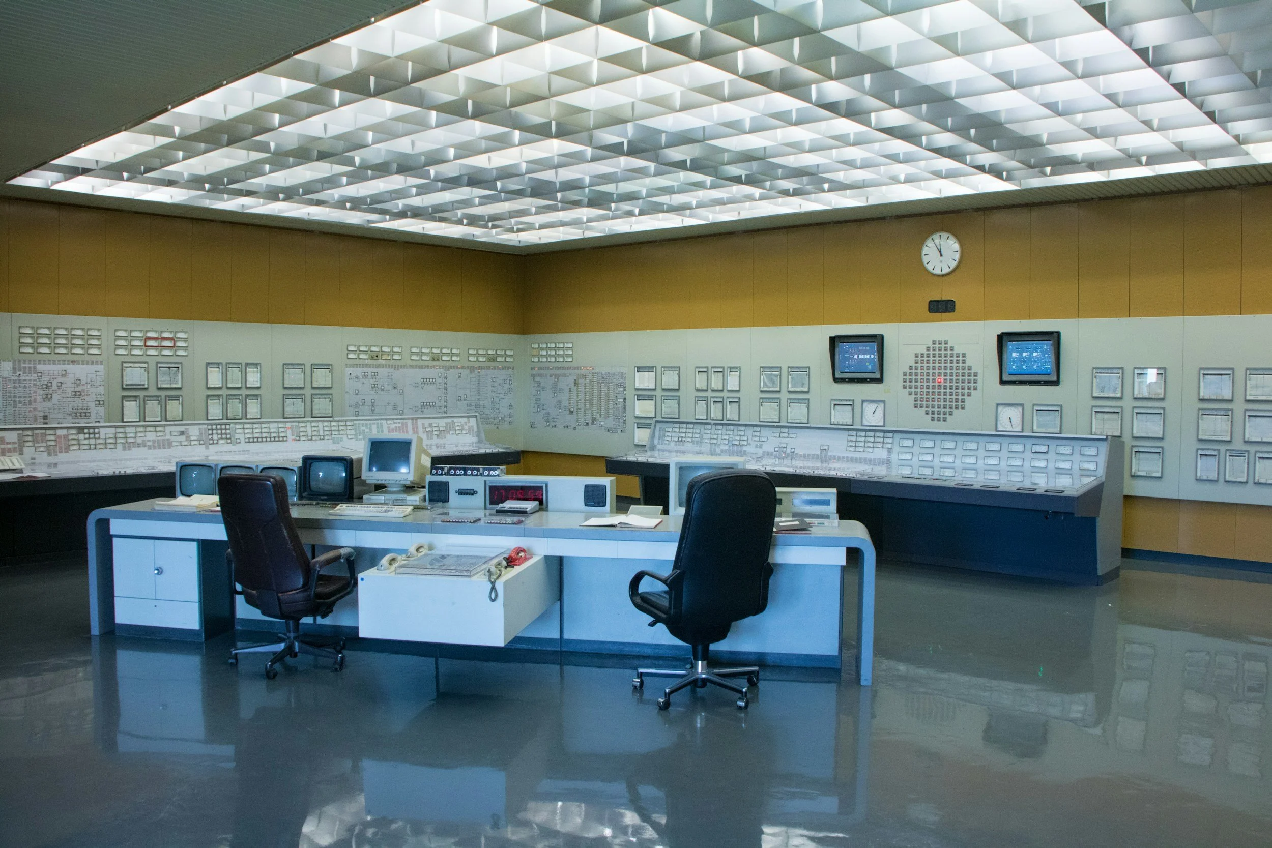 A large control room with multiple control panels, a central desk with old computer monitors, chairs, and wall-mounted screens, clocks, and diagrams.