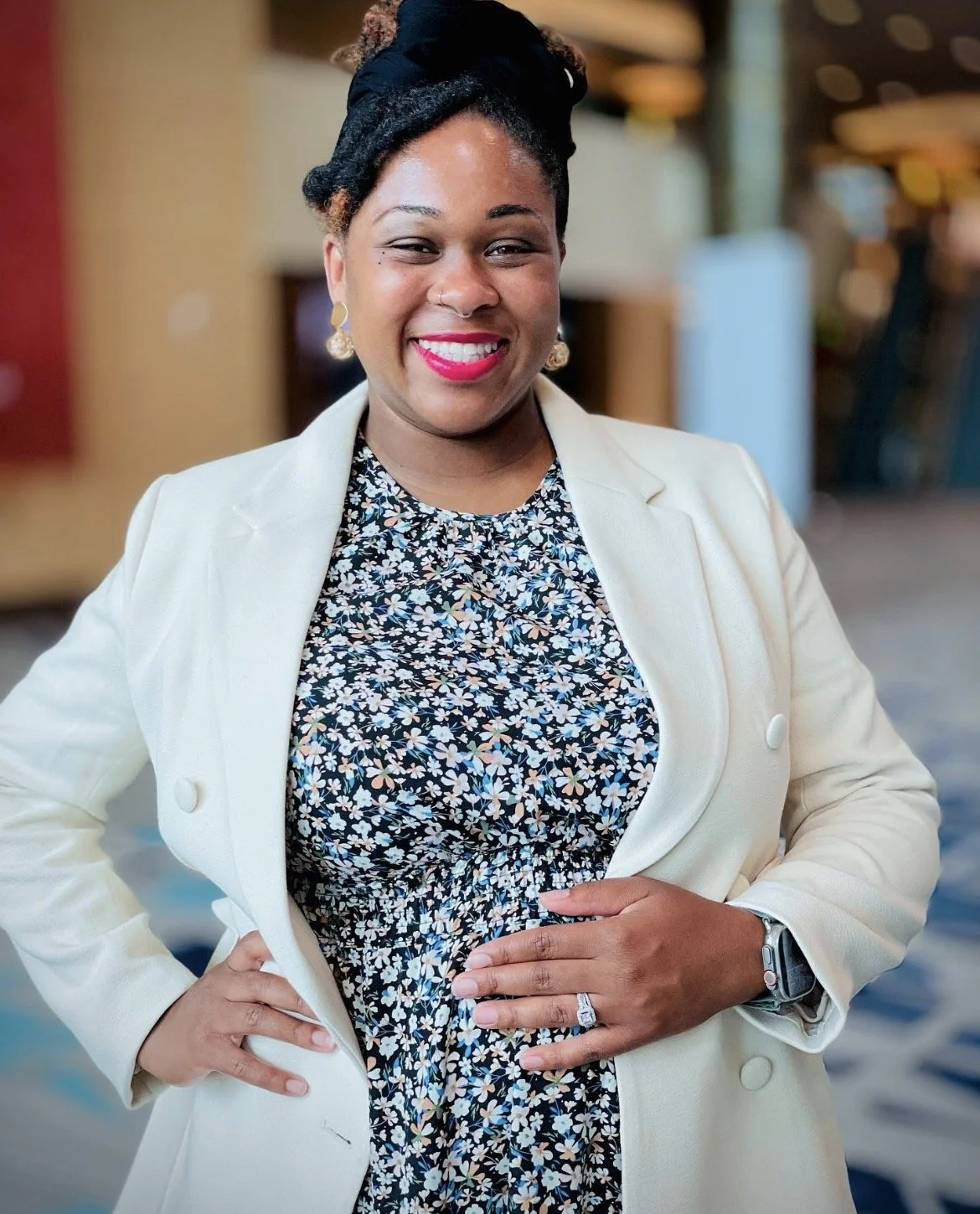 A woman with dark skin and curly hair styled in an updo, wearing a white blazer over a floral dress, smiling at the camera in an indoor setting.