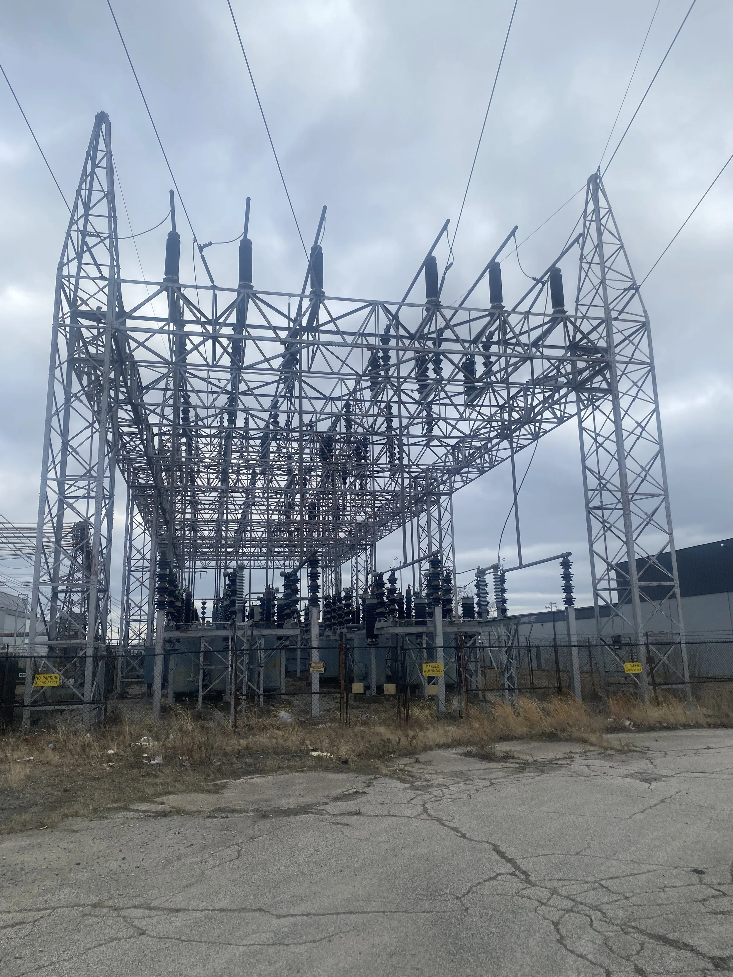 An electrical substation with metal framework, insulators, and electrical equipment, surrounded by fencing, under a cloudy sky.