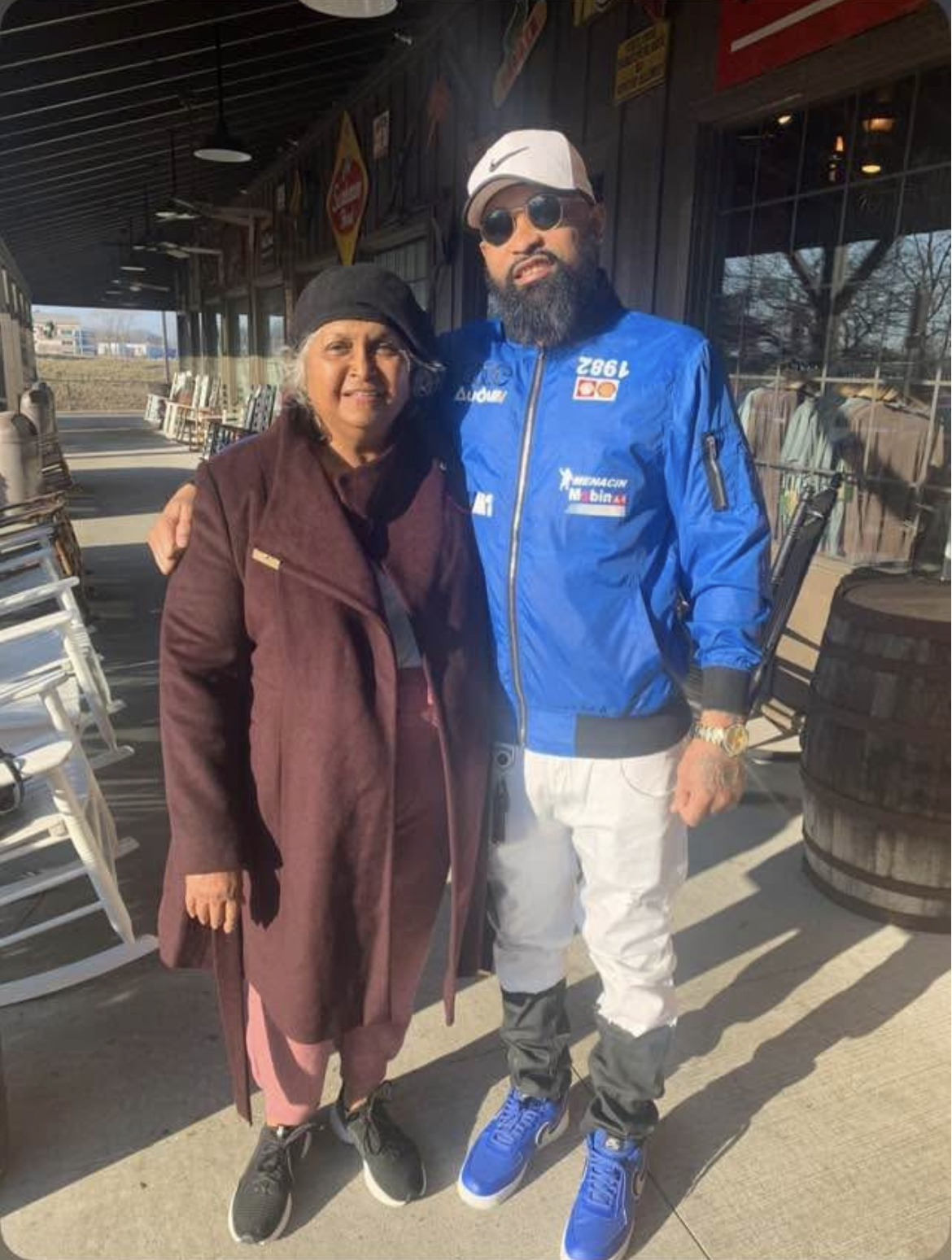 Two people standing outside at a restaurant or cafe, posing for the photo. The woman on the left has gray hair, is wearing a black hat, maroon coat, and black shoes. The man on the right has a beard, is wearing sunglasses, a white cap, a blue racing jacket with sponsor logos, white pants, and blue shoes.