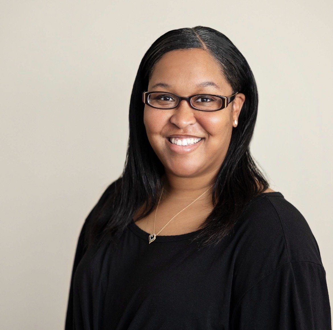 A smiling woman with glasses, wearing a black top, necklace, and earrings, posed against a plain light background.