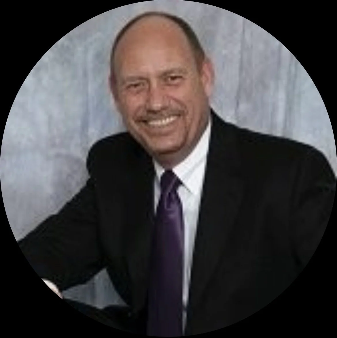 A man in a black suit, white shirt, and purple tie smiling in front of a light-colored background.