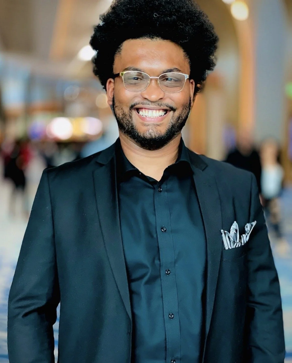 A smiling man with glasses and a beard wearing a black suit and shirt, standing in a well-lit indoor setting with blurred background of people and lights.