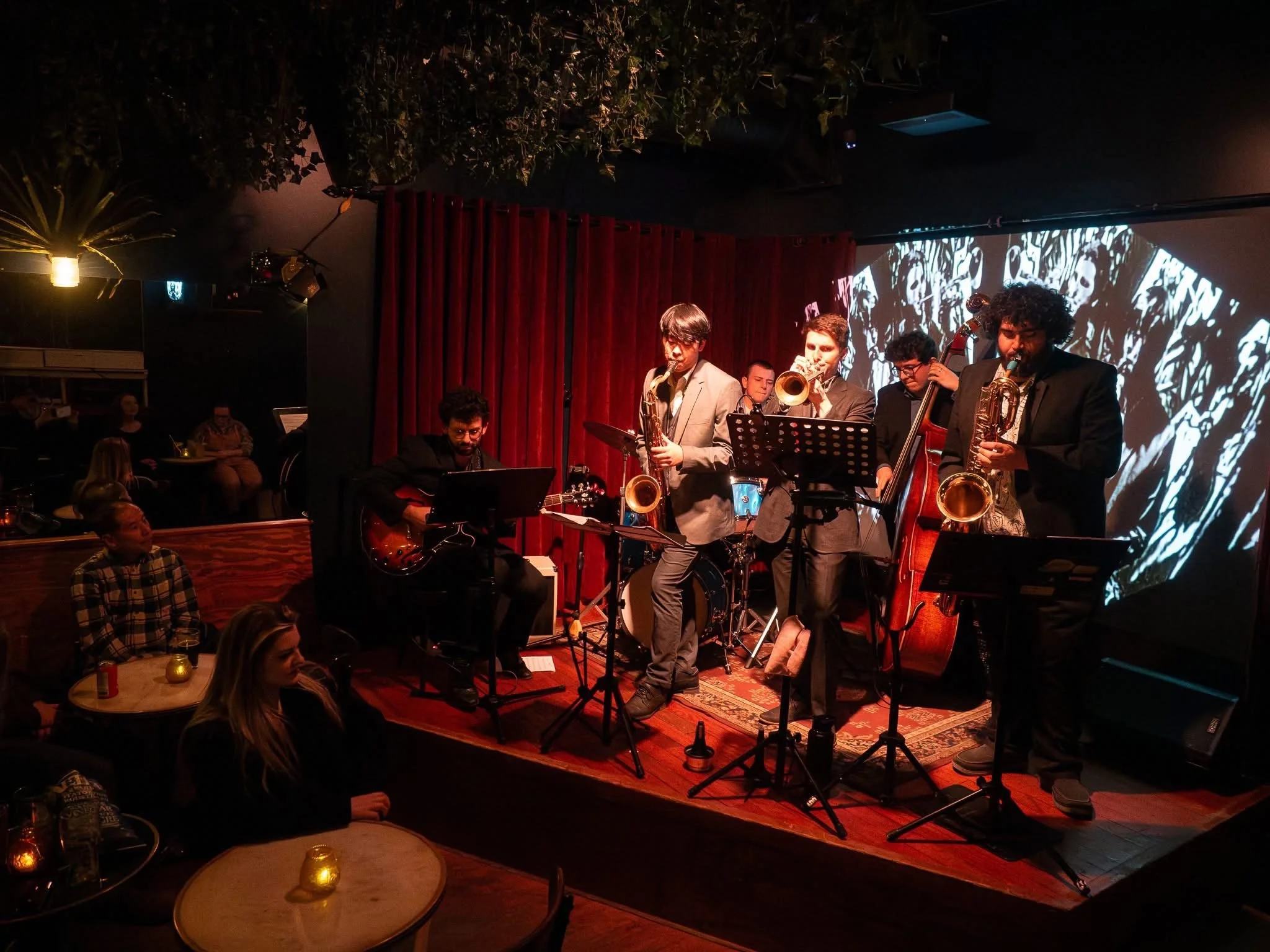 A jazz band performs on a small stage in a dimly lit venue with a red curtain backdrop and a black-and-white graphic screen. The band includes a guitarist, two saxophonists, a trombone player, a double bassist, and a drummer. Audience members sit at small tables with lit candles.