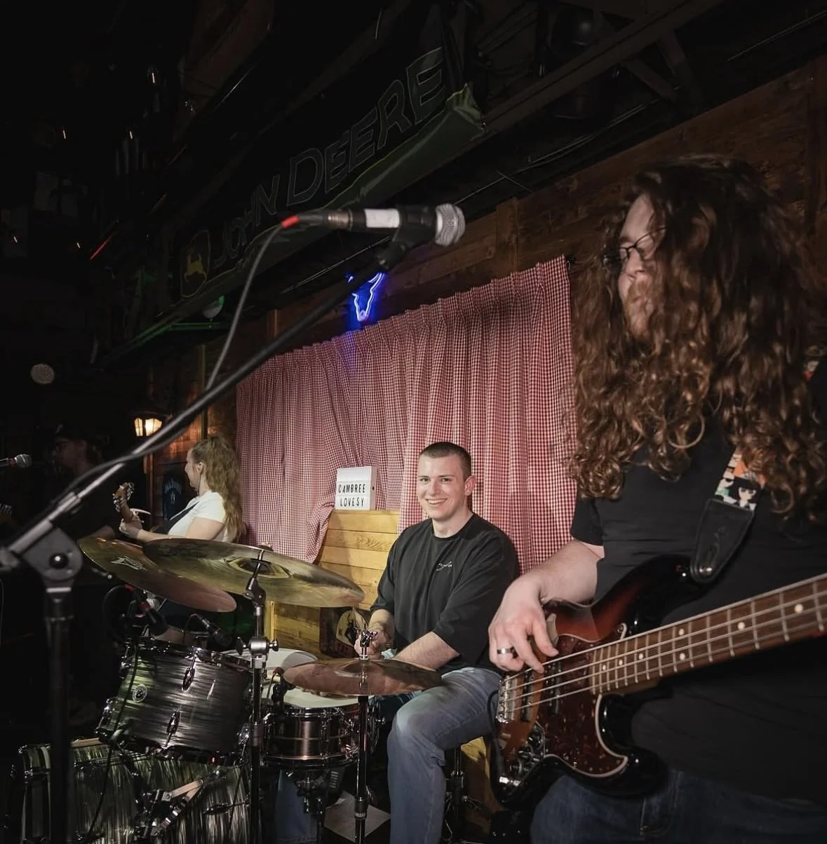 A band performing live on stage, including a drummer and a guitarist. The drummer is smiling at the camera, and the guitarist has long curly hair and glasses. The stage has a red checkered curtain backdrop and a sign that reads 'CAMBREE LOVESY'.