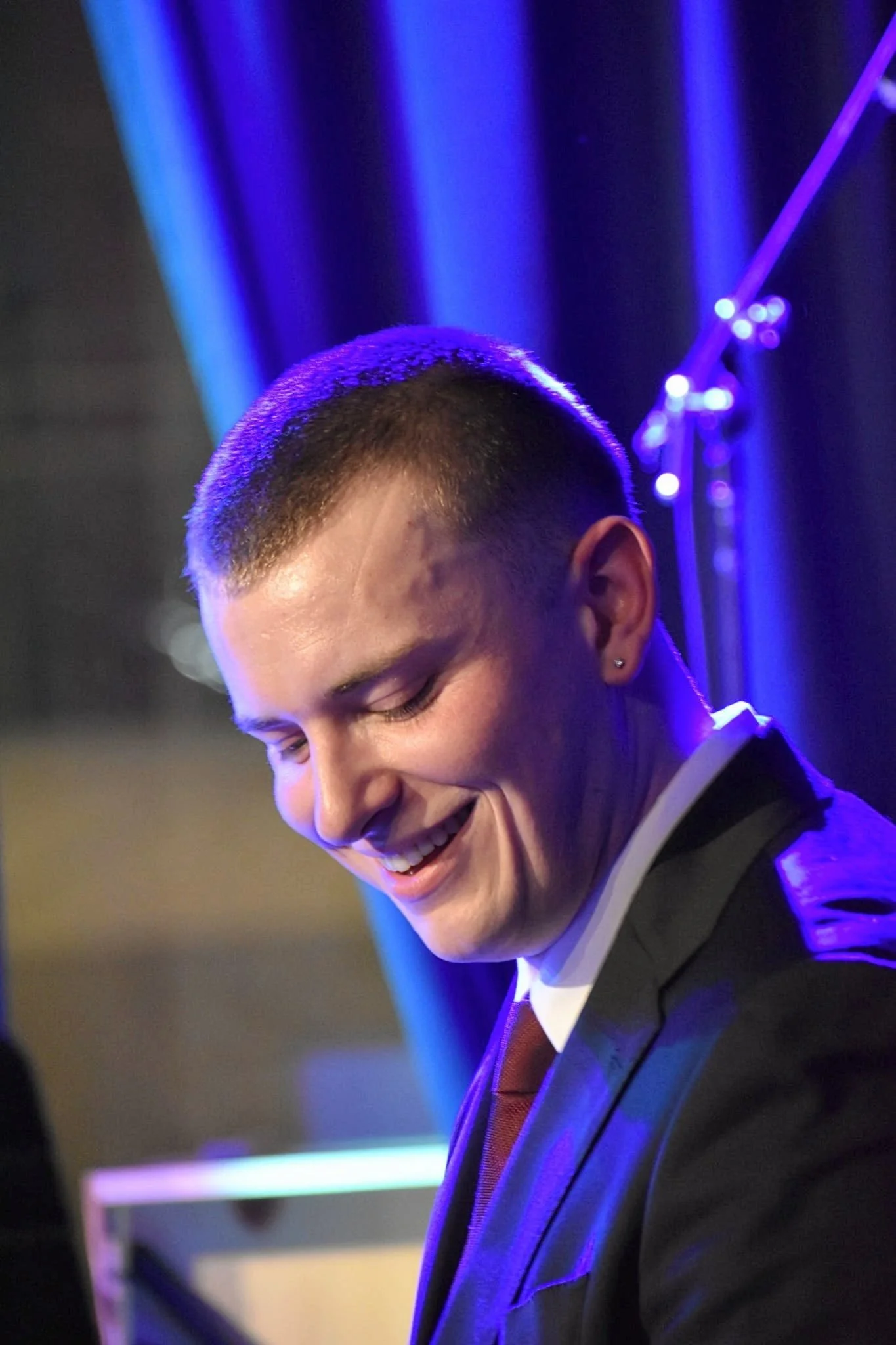 A young man smiling and looking down, dressed in a suit, with a dark background and blue lighting.