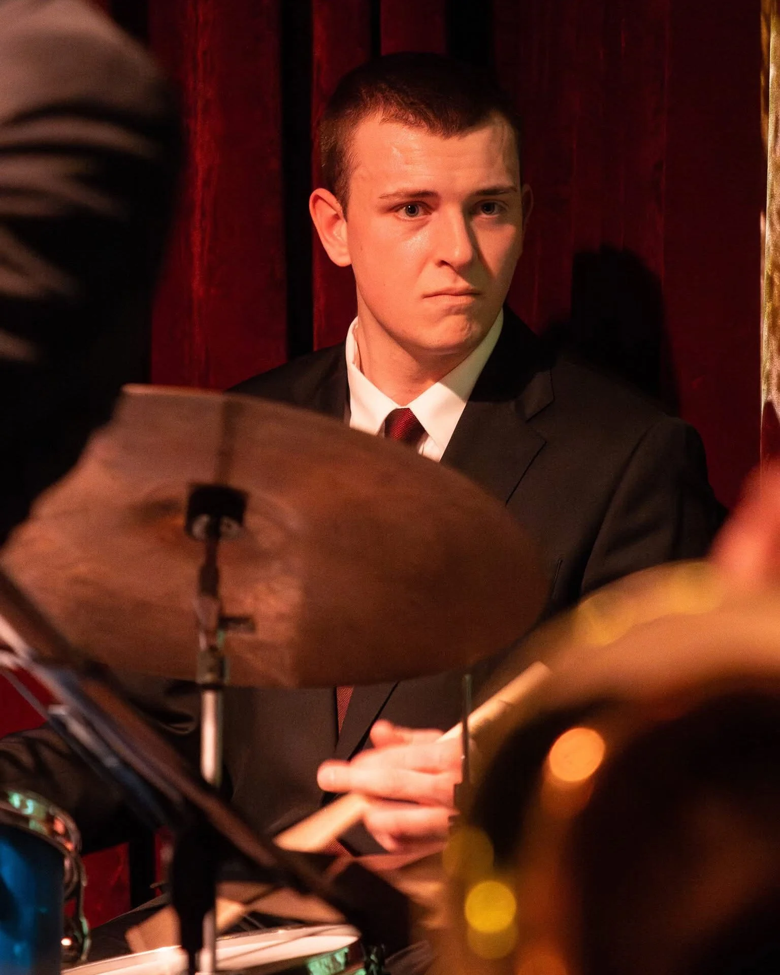 A young man in a tuxedo sitting behind a drum set, looking intently to his right at a jazz band performance.