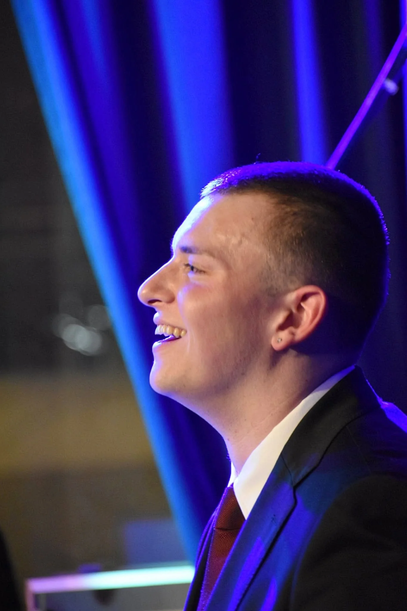 A young man with short hair and earrings, dressed in a suit and tie, smiling in profile against a background with blue and purple stage lighting.