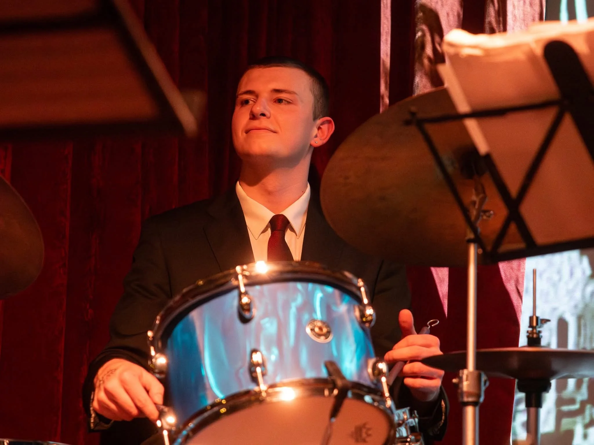 Young man in a black suit and white shirt, playing drums in a dimly lit room with red curtains.
