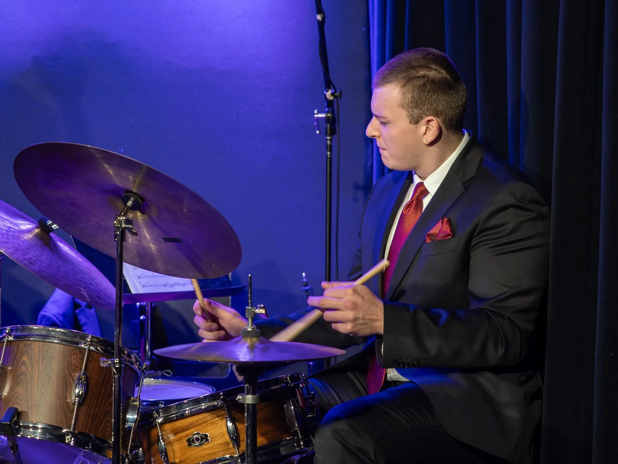 A man in a black suit and red tie playing a drum set on stage with dark curtains behind him.