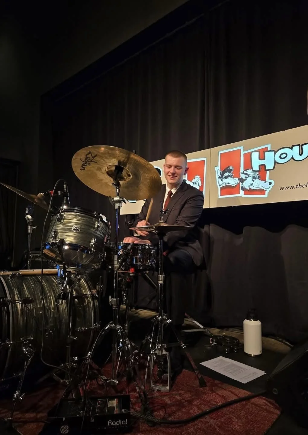A man in a suit playing drums on stage at a comedy show.