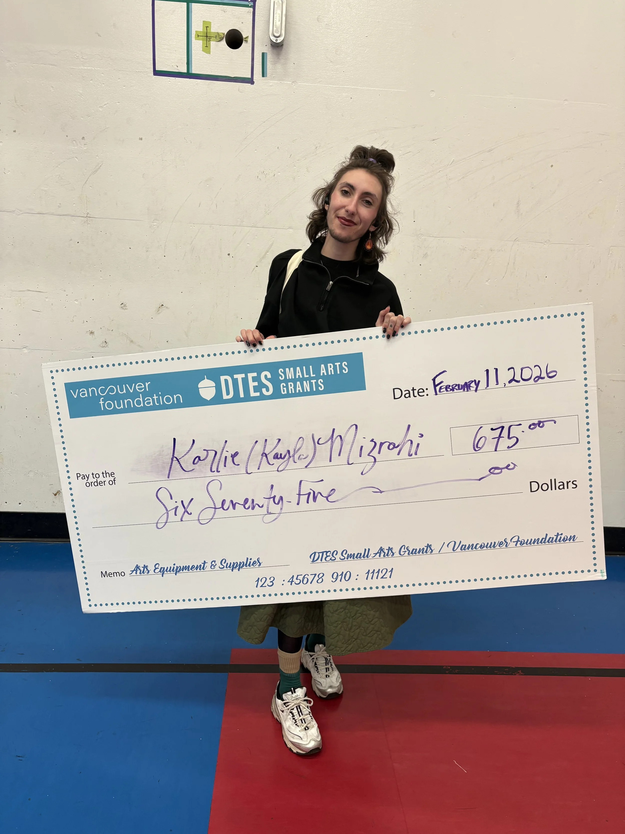 Karlie Mizrahi in a Black 3/4 Zip with a Black Turtleneck and their Gold and Silver Chain with a Camoflauge Green Skirt, Green Socks with Gold Rims, Black Tights and Black, Silver and White Sneakers. They are holding a check for 675.00