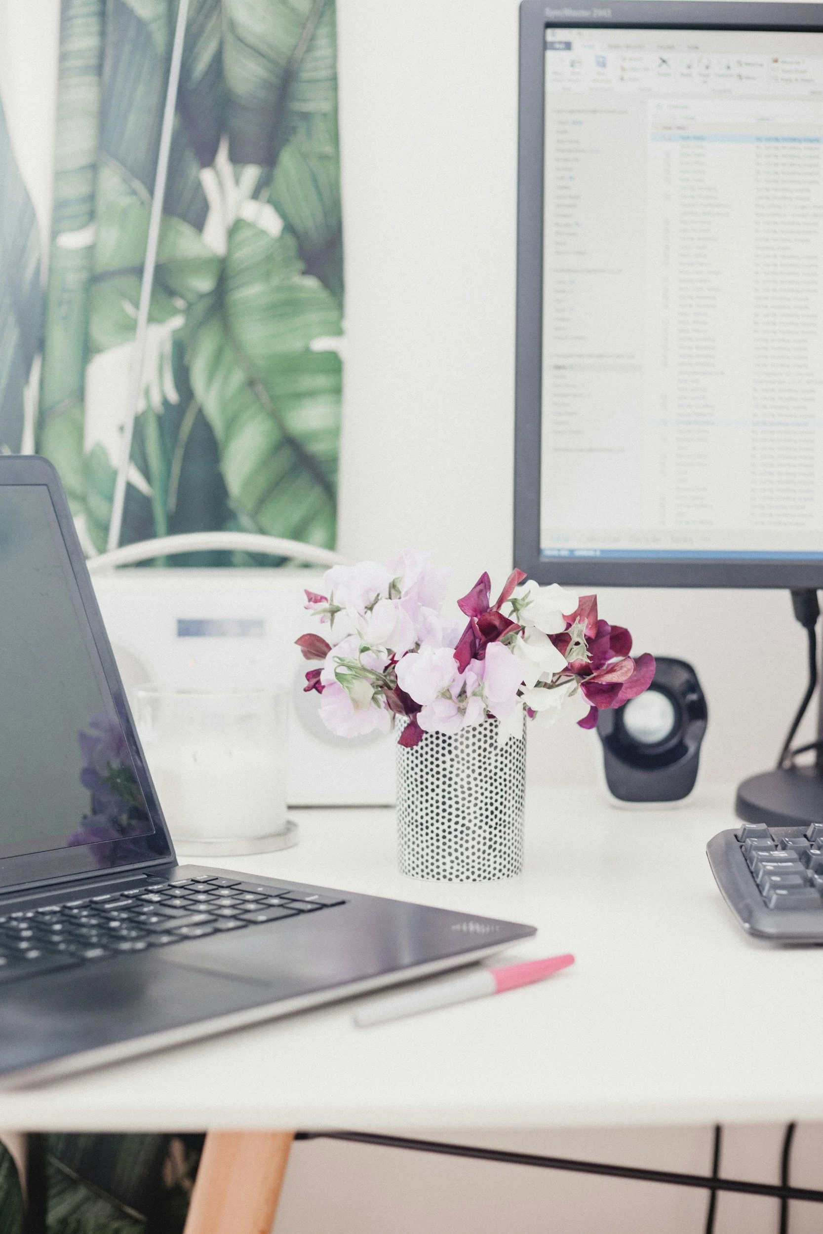 Medical reception in a private rooms with an open laptop, a pink marker, a small potted plant with pink and white flowers, a computer monitor displaying an email inbox, a black speaker, a computer keyboard, .