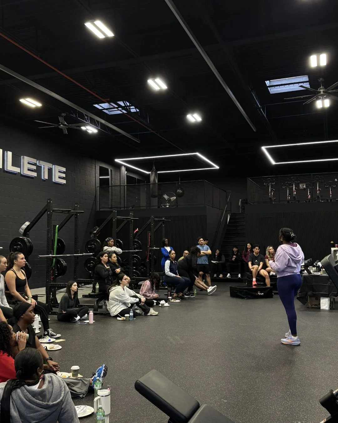 A group of people in a gym watching a fitness instructor. The gym has black walls, weight racks on the side, and bright ceiling lights. Some people are seated on the floor, others on benches, with water bottles and snacks nearby.