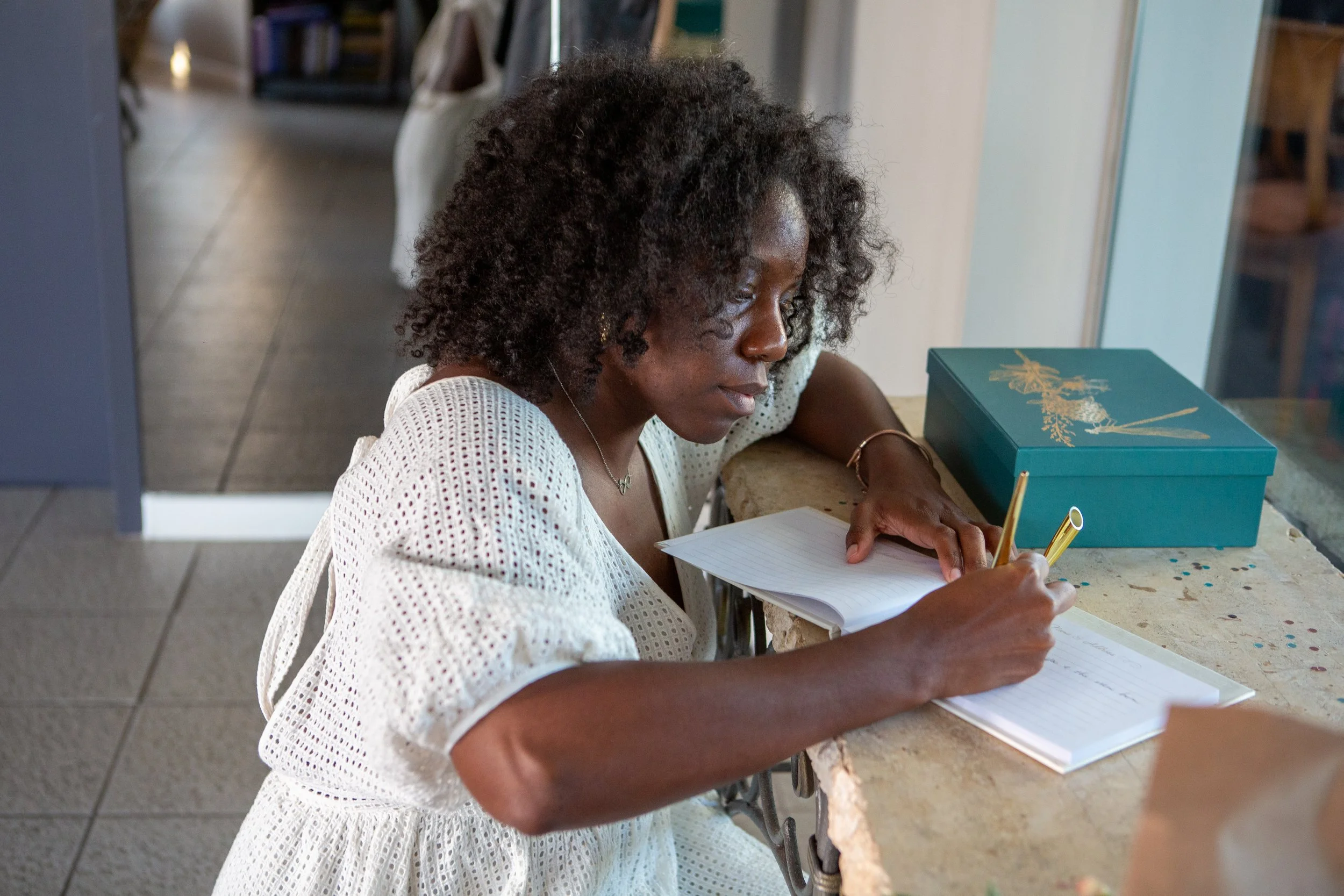 Woman with curly hair writing in a notebook at a cafe counter, with a teal box nearby.