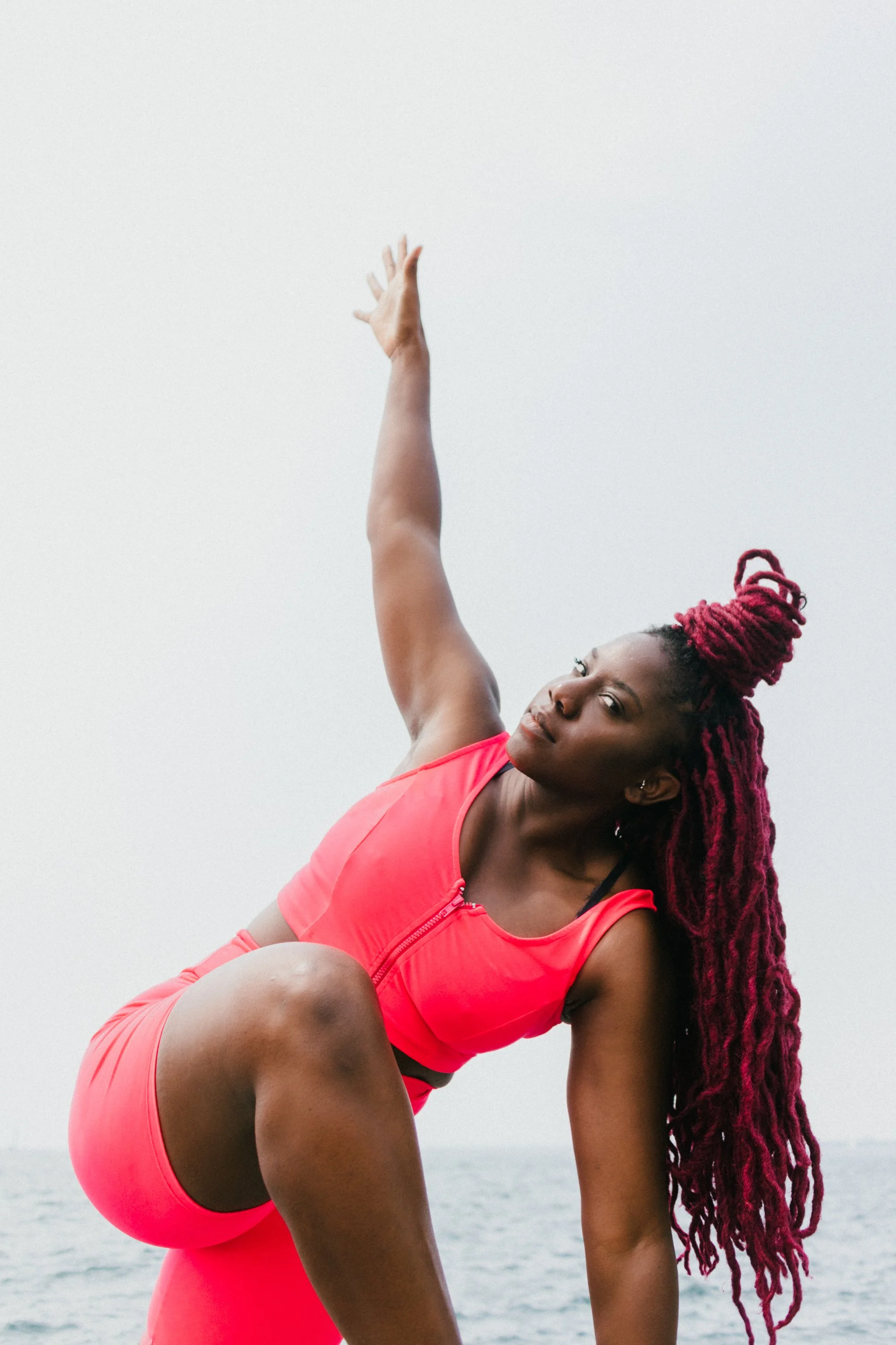 A woman practicing yoga outdoors near water, wearing a bright pink sports bra and matching leggings, with long braided hair in a bun.