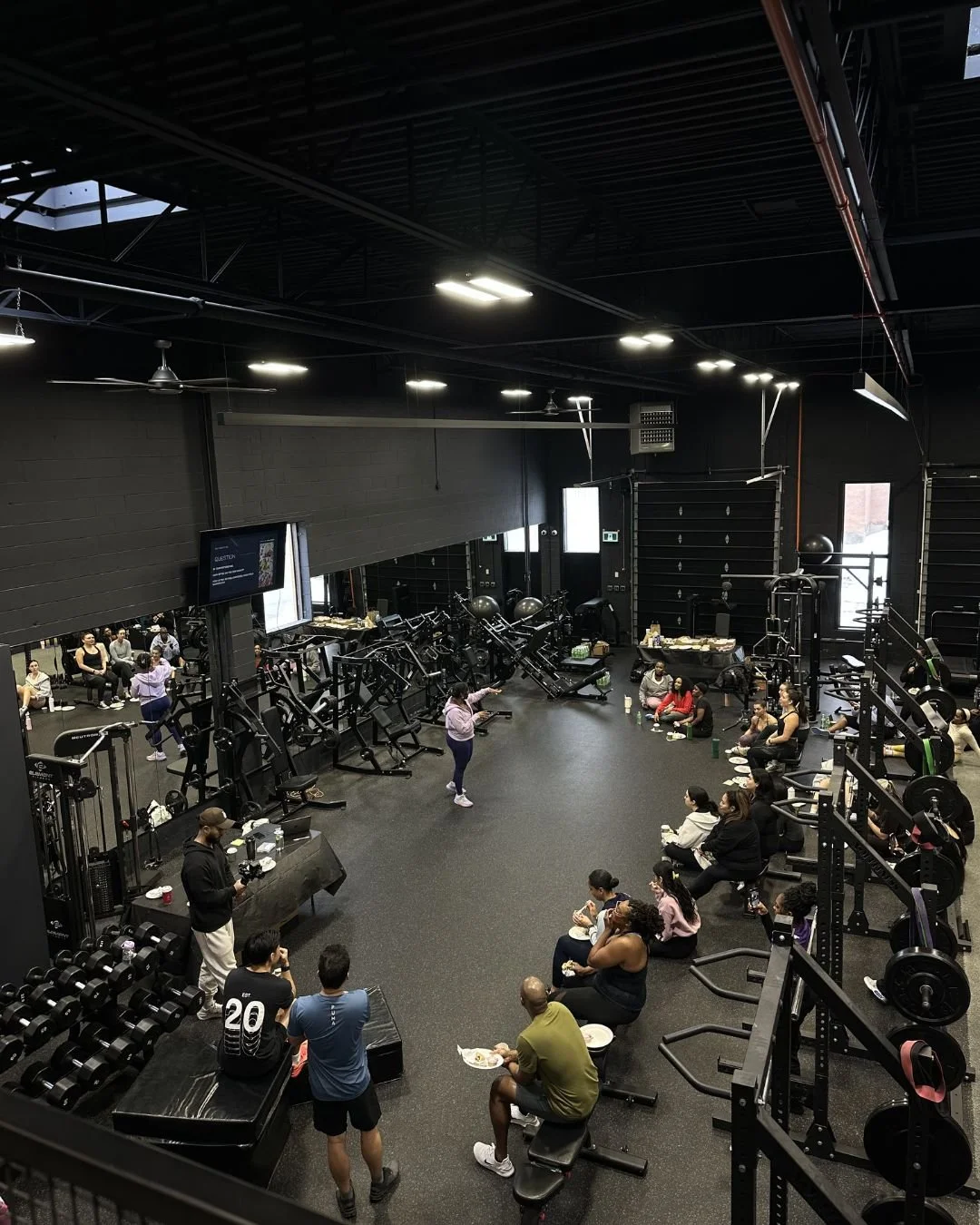 People sitting on benches in a gym during a fitness class, with workout equipment like dumbbells and machines visible, and an instructor at the front leading the class.