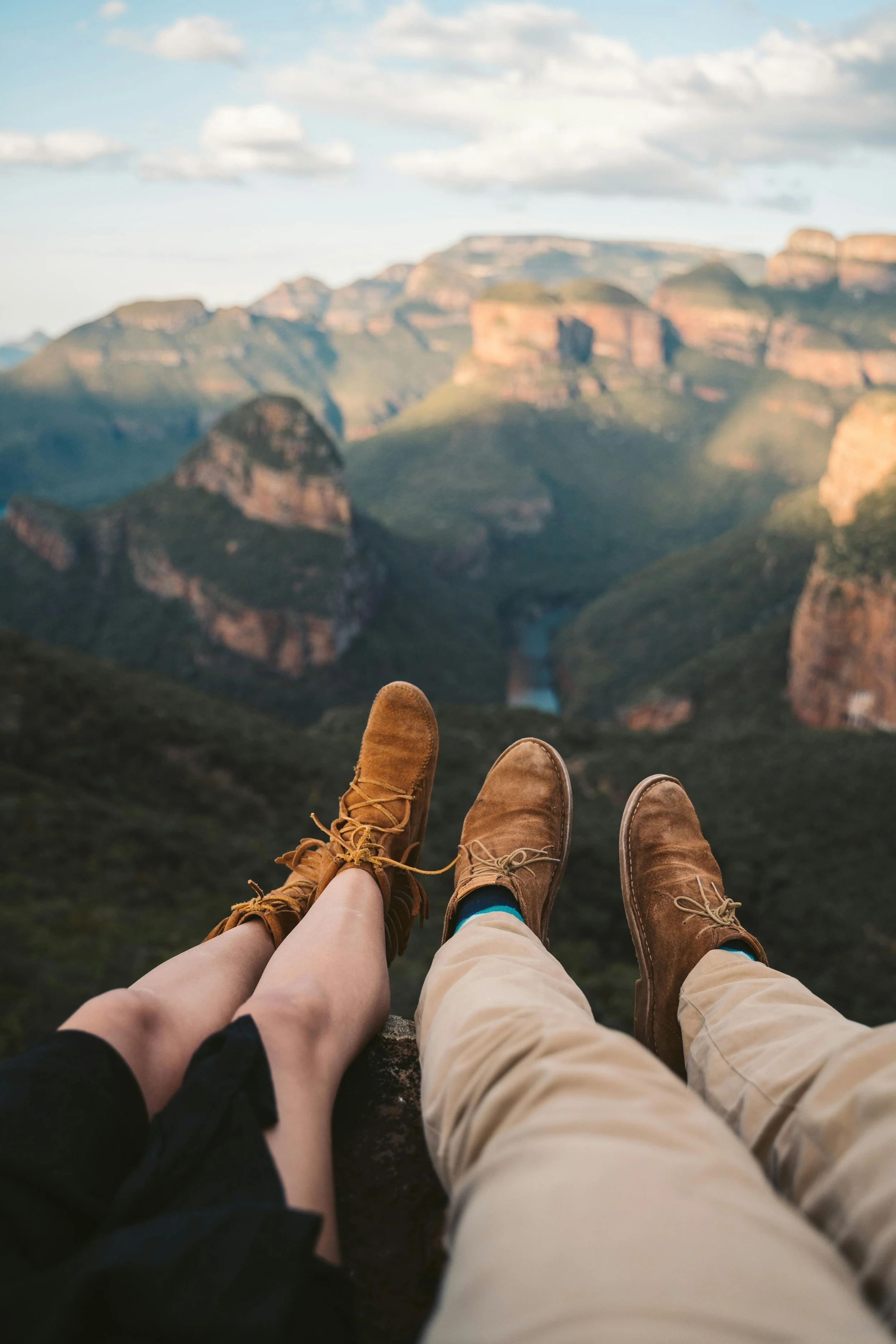 A couple sitting on a ledge with their legs extended, overlooking a scenic canyon with cliffs and a river below, under a partly cloudy sky.