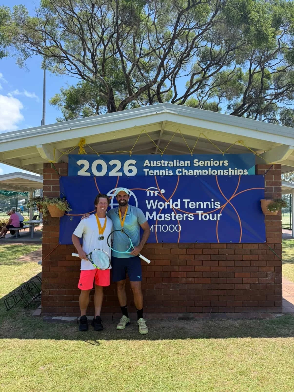 Two tennis players standing together with medals around their necks and tennis rackets, smiling in front of a sign for the 2026 Australian Seniors Tennis Championships and World Tennis Masters Tour held outdoors under a pavilion, with trees and a bright sky in the background.
