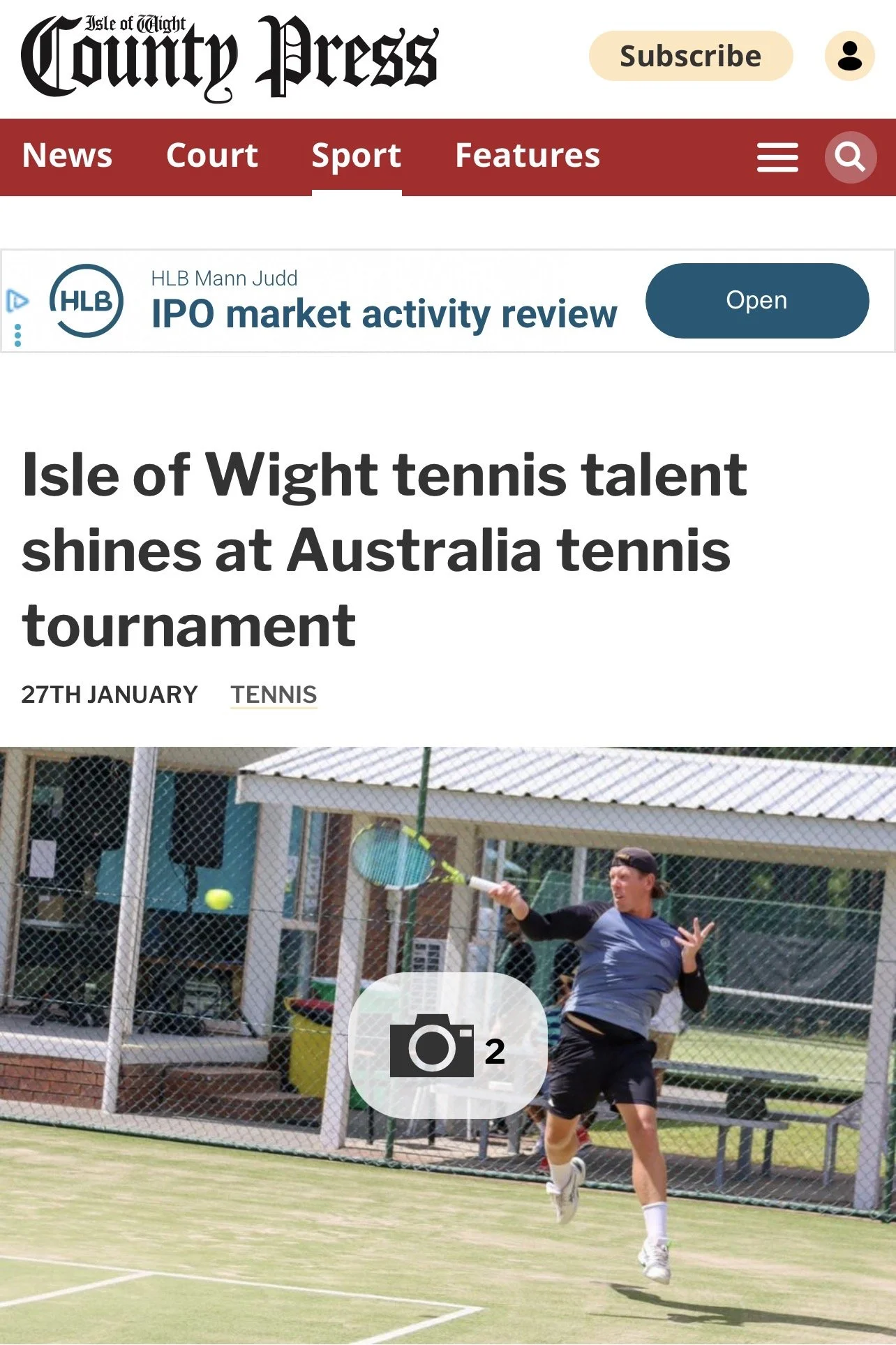 A male tennis player in mid-air hitting a tennis ball with a racket on an outdoor tennis court, with a fence and a building in the background.