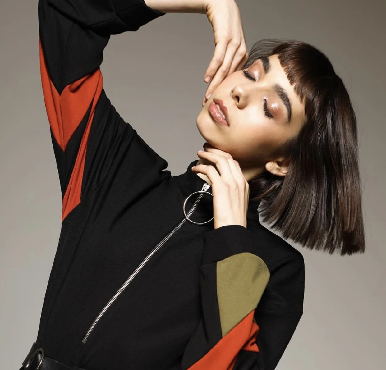 A woman with shoulder-length dark brown hair and bangs poses with eyes closed, wearing a black jacket with red and green accents, and touching her face gracefully.