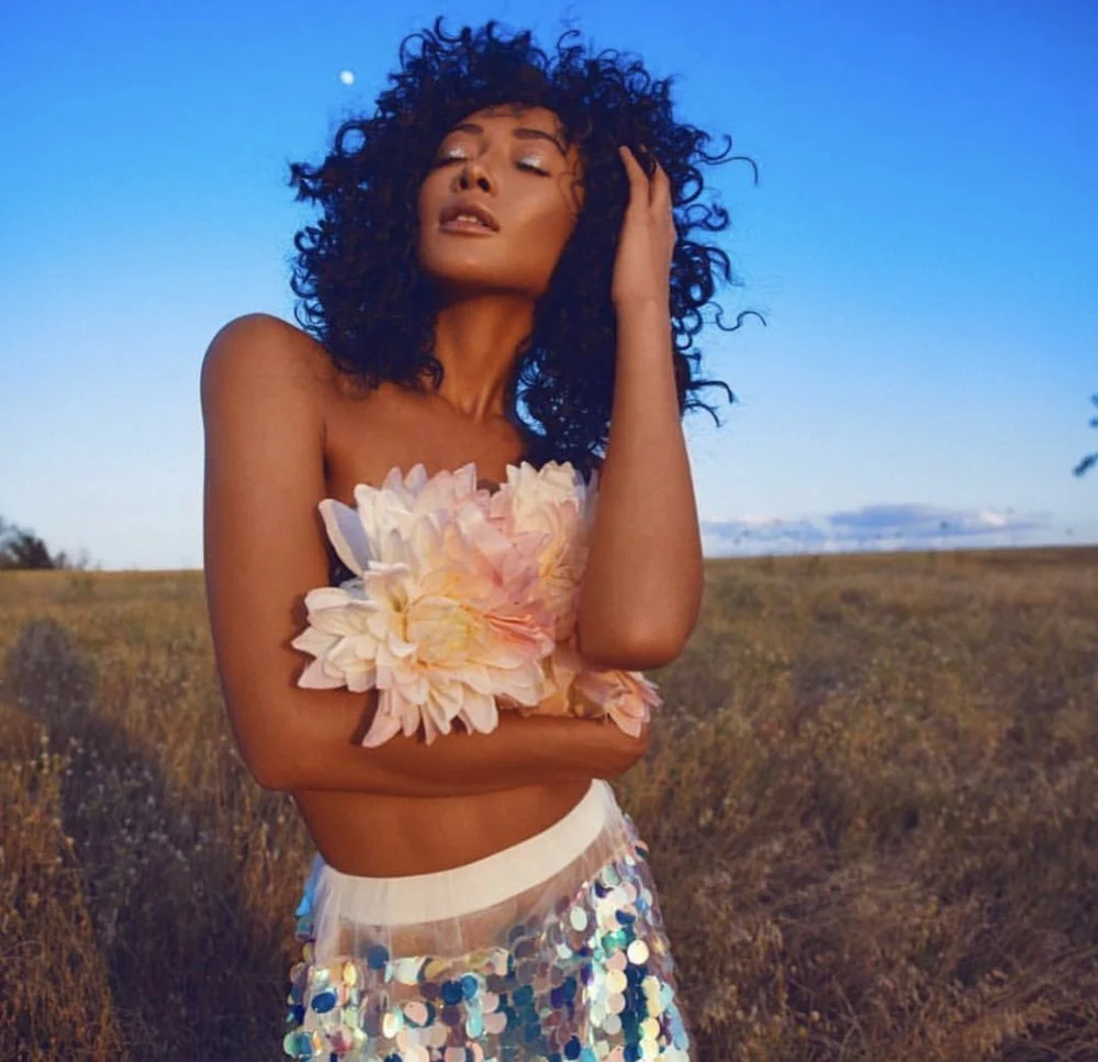 A woman with curly black hair and tan skin stands outdoors in a field during sunset, holding a bunch of large pink and white flowers close to her chest with one arm, and touching her hair with the other. She wears a scarf covered in shiny, silver seq