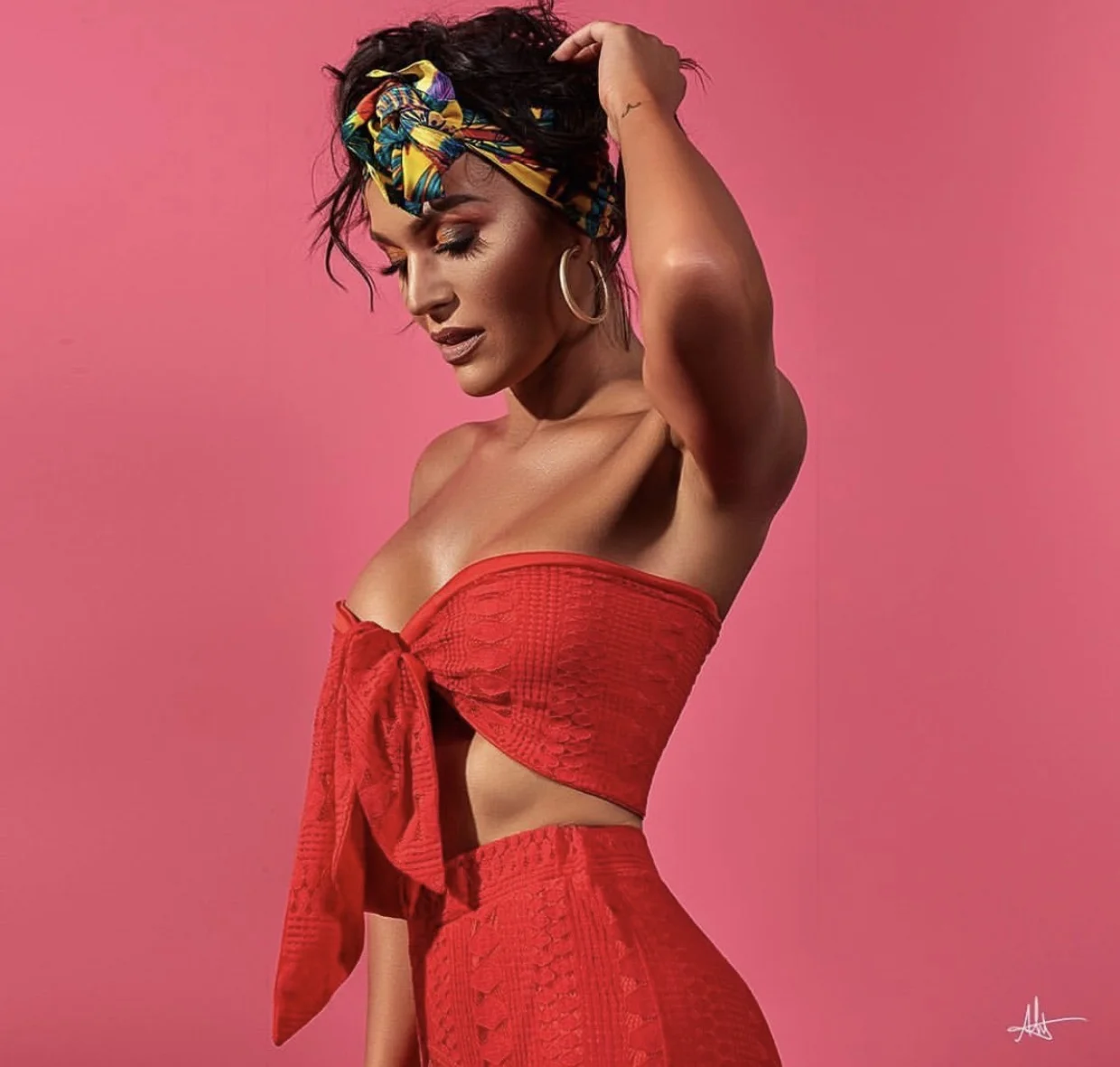 A woman with dark hair and large hoop earrings poses against a pink background. She is wearing a colorful headscarf and a red crop top with a bow in the front, matching high-waisted shorts, and is touching her head with one hand.