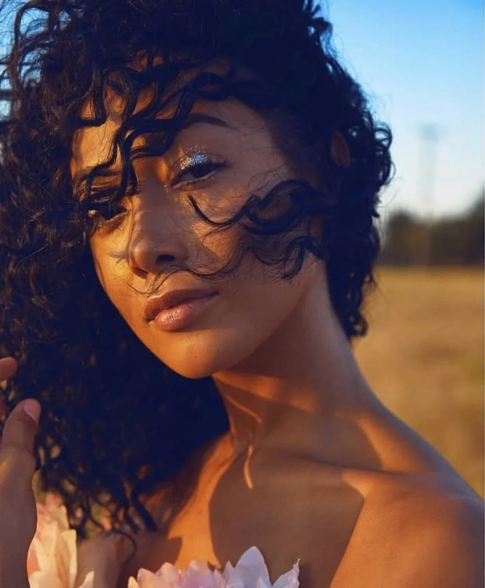 A woman with curly dark hair outdoors during sunset, holding a pink flower, with her face partially shadowed and a serene expression.