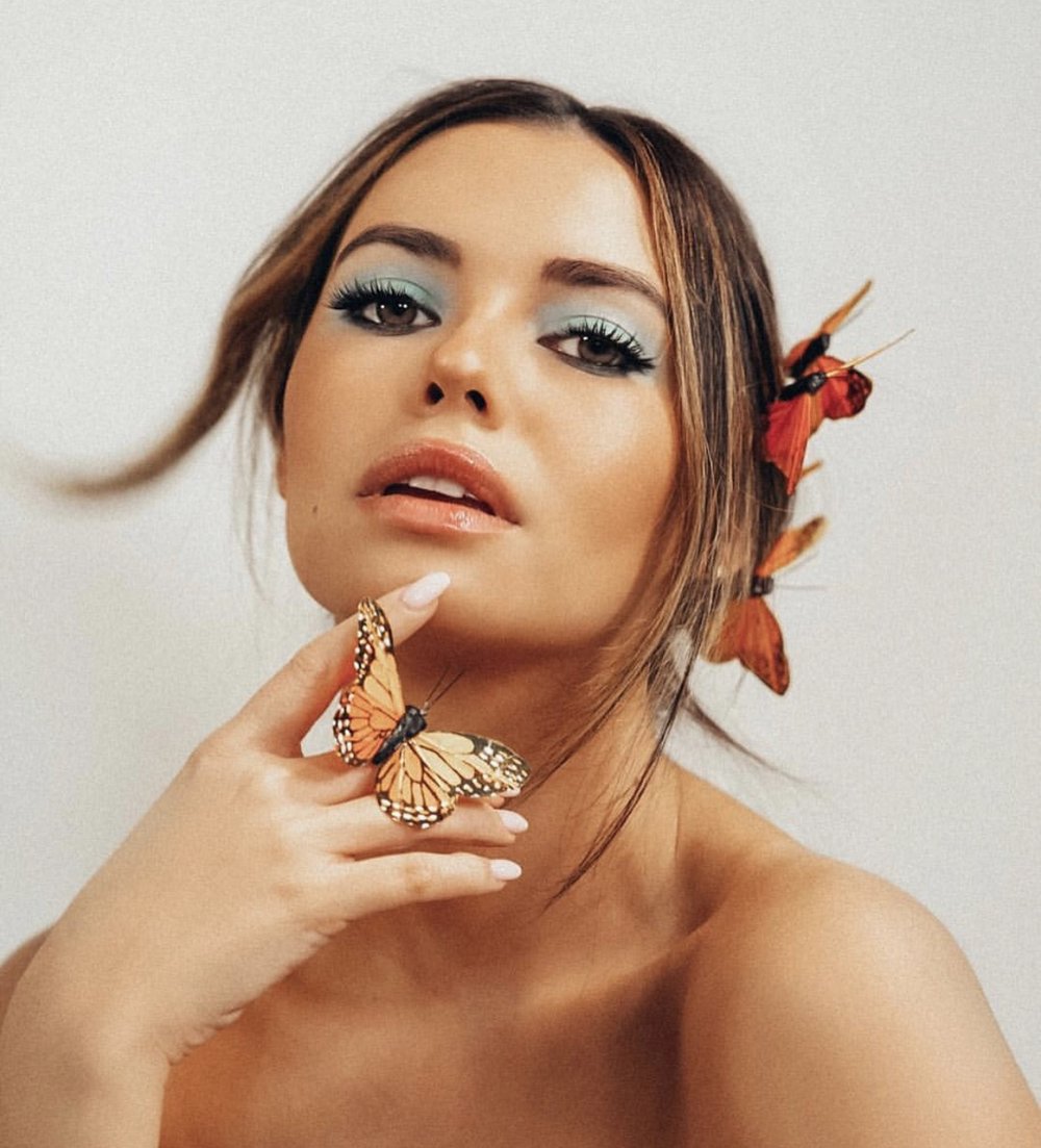 A woman with shoulder-length brown hair, wearing light blue eyeshadow, has butterflies on her face and hand, standing against a plain background.