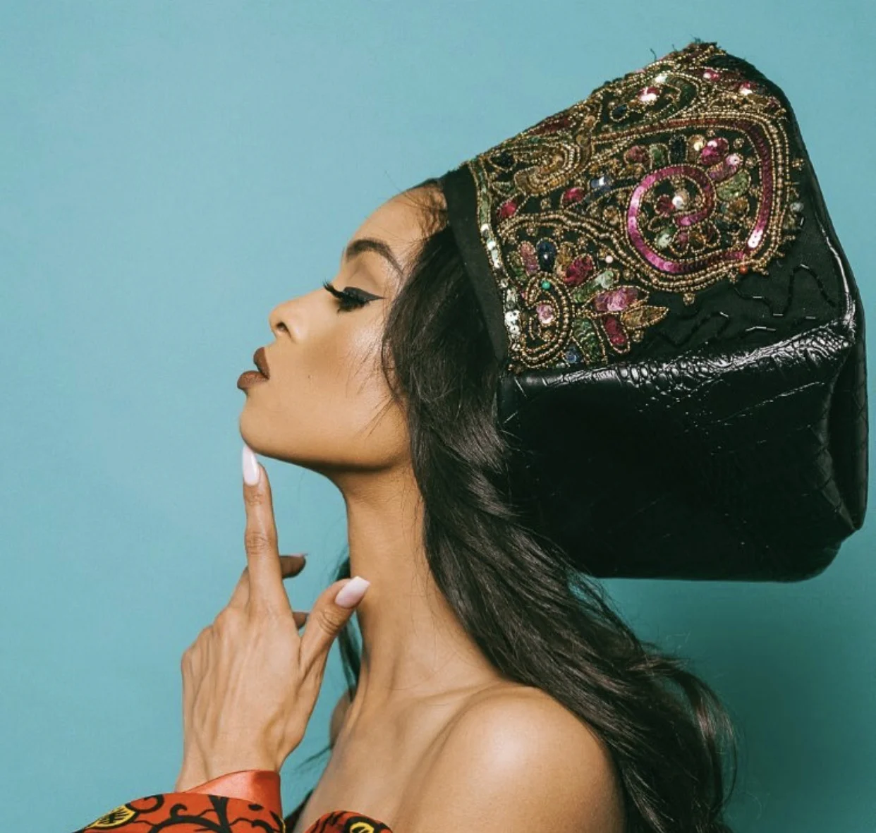 A woman with long black hair and makeup, wearing a large, black hat with colorful embroidery and beads, touching her lips with her finger against a light blue background.