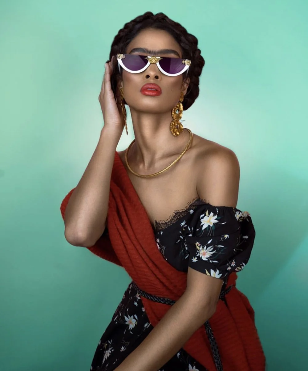 Woman with dark hair in braids wearing large purple sunglasses, gold jewelry, a black floral off-shoulder dress, and a red shawl, against a gradient green background.