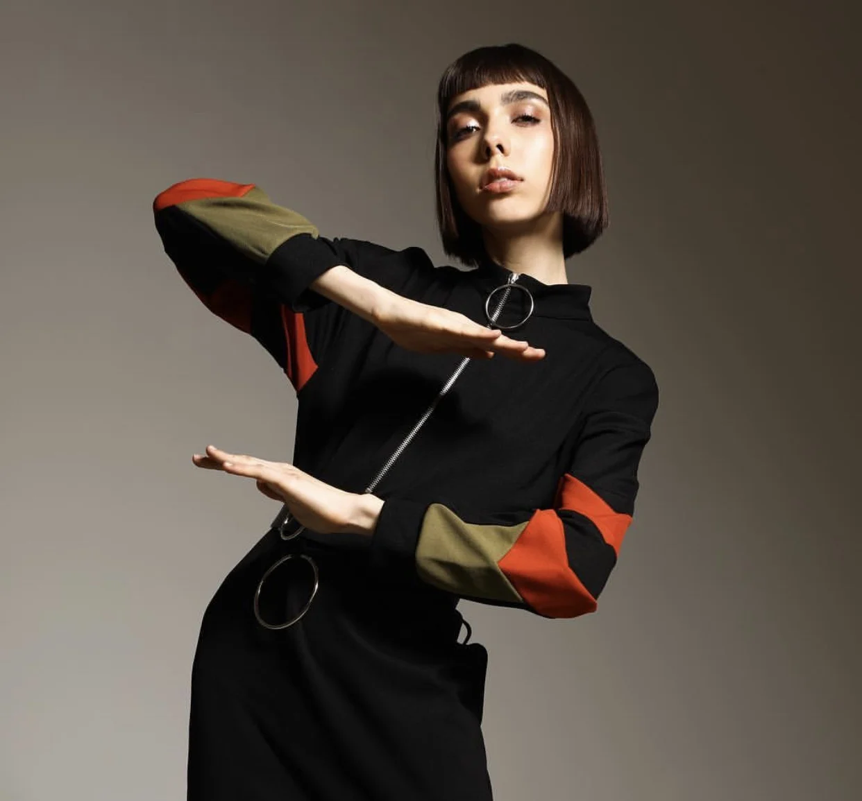 A woman with short brown hair wearing a black dress with colored patches on the sleeves and a metal ring zipper pull, standing against a plain background.