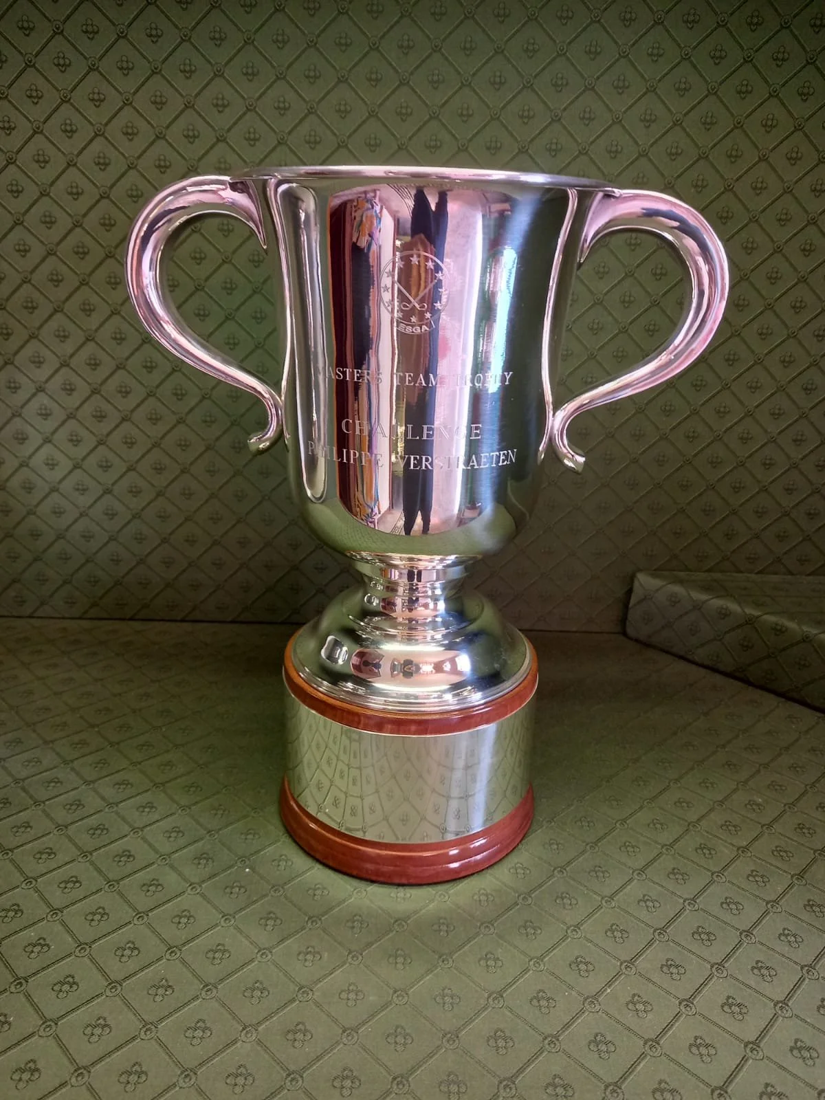 A silver trophy cup with handles sits on a polished base with wood trim. The cup's surface is reflective, showing a person taking the photo and a room with patterned wallpaper and furniture. The cup has engraved text and a logo.