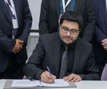 Man in gray suit signing a document at a table, surrounded by several people in business attire.