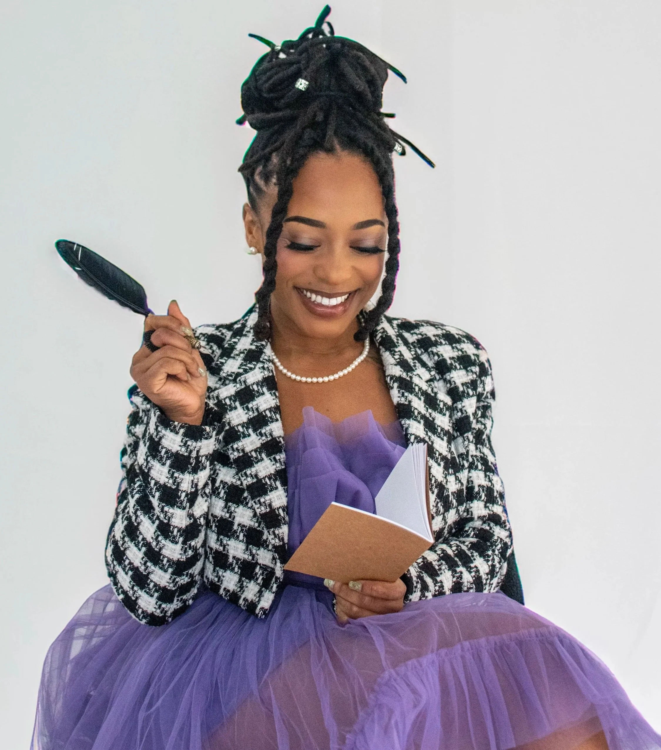 A smiling woman with dreadlocked hair styled in a high bun, wearing a checkered blazer, pearl necklace, and pearl earrings, holding a bouquet of purple flowers and a brown envelope, standing against a plain white background.