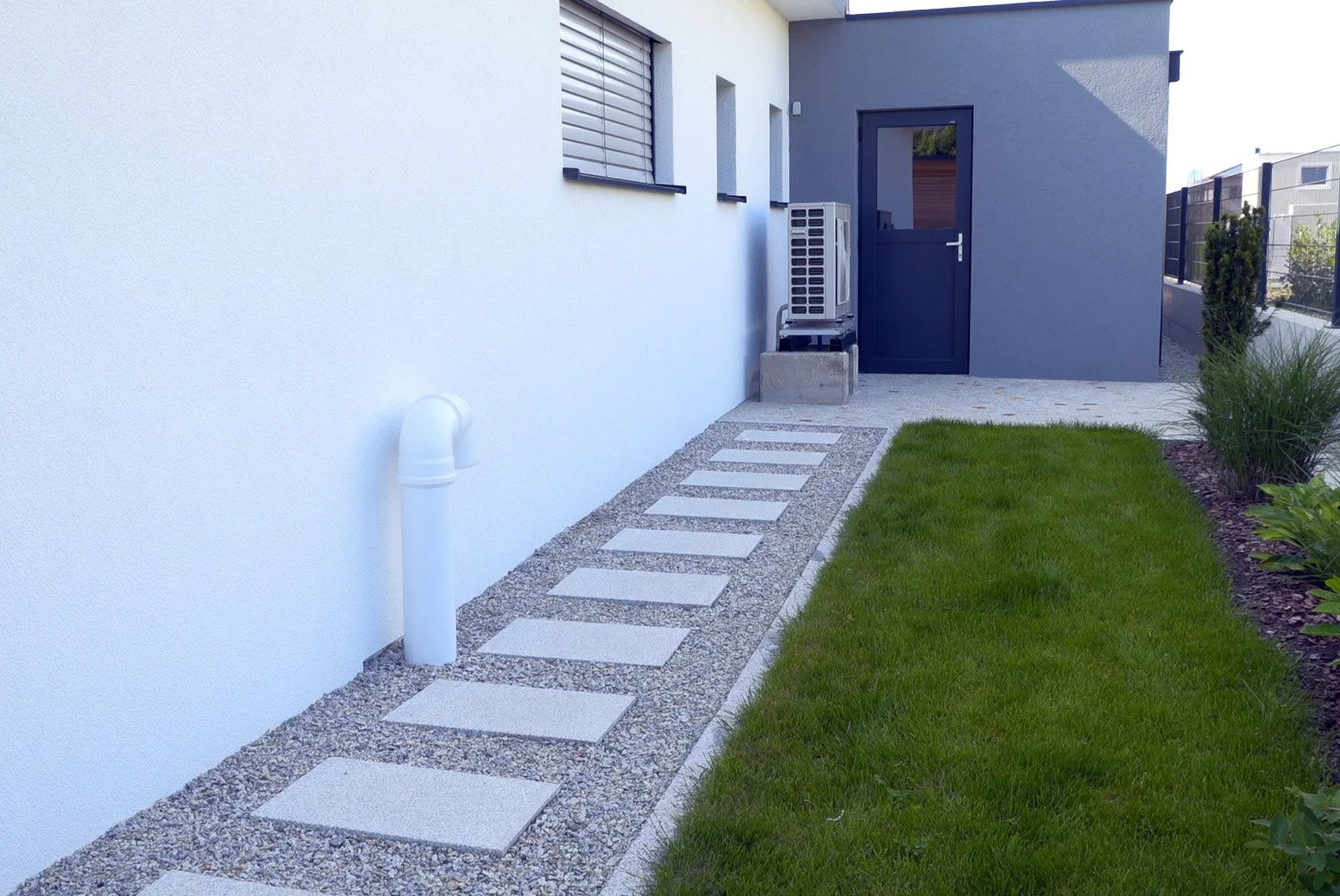 Gartenweg mit Gehwegplatten, Rasen und Büsche vor modernen Haus mit grau-weißer Wand und dunkler Tür.