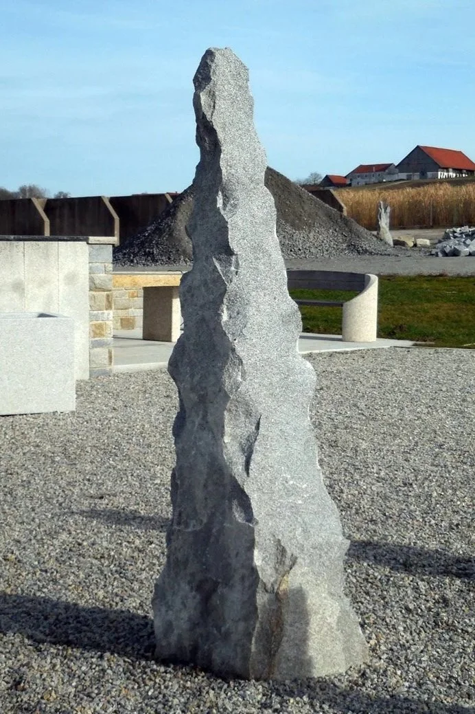 Zwei Fotos von vertikalen Naturstein-Skulpturen im Freien, eine große und rauhe mit unregelmäßigen Oberflächen, die andere kleiner und mit einer glatten Seite, umgeben von Steinhaufen und Bauwerken.