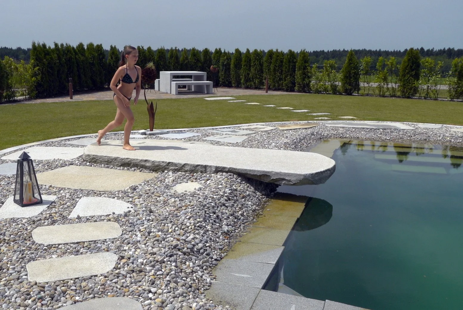 Ein Mädchen in Bademode läuft über einen Steinweg, der zu einem Swimmingpool führt, im Garten mit Bäumen im Hintergrund, sonniges Wetter.