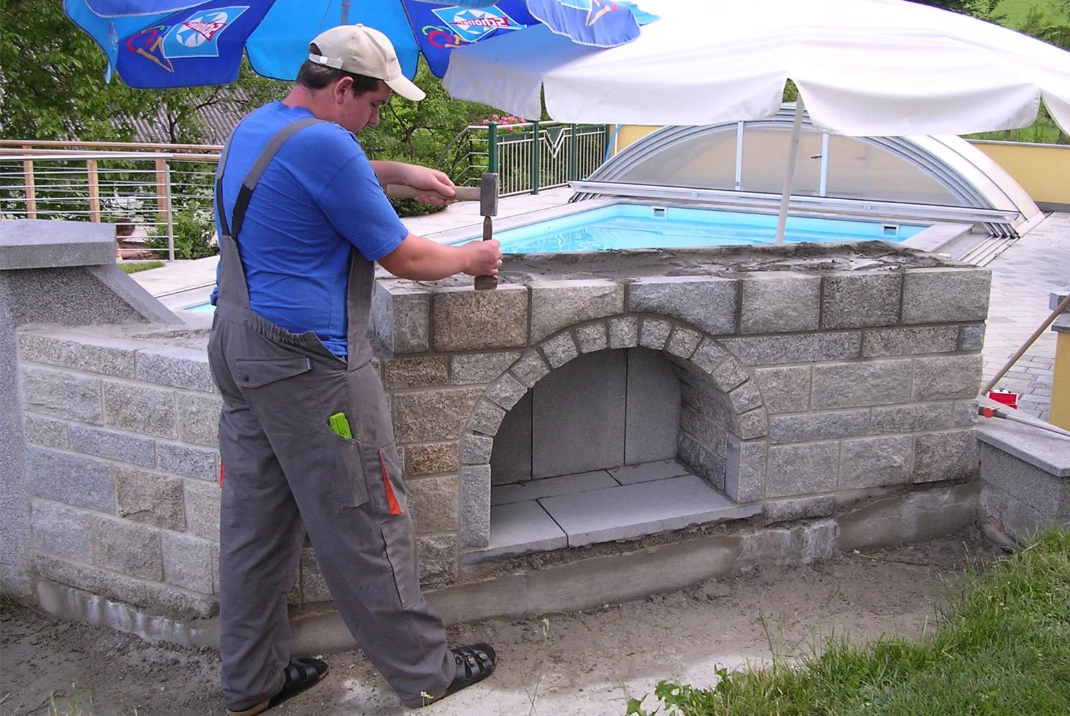 Ein Mann arbeitet an einem neuen Außenkamin oder Grill im Garten, mit einem Pool im Hintergrund, bei bewölktem Himmel.