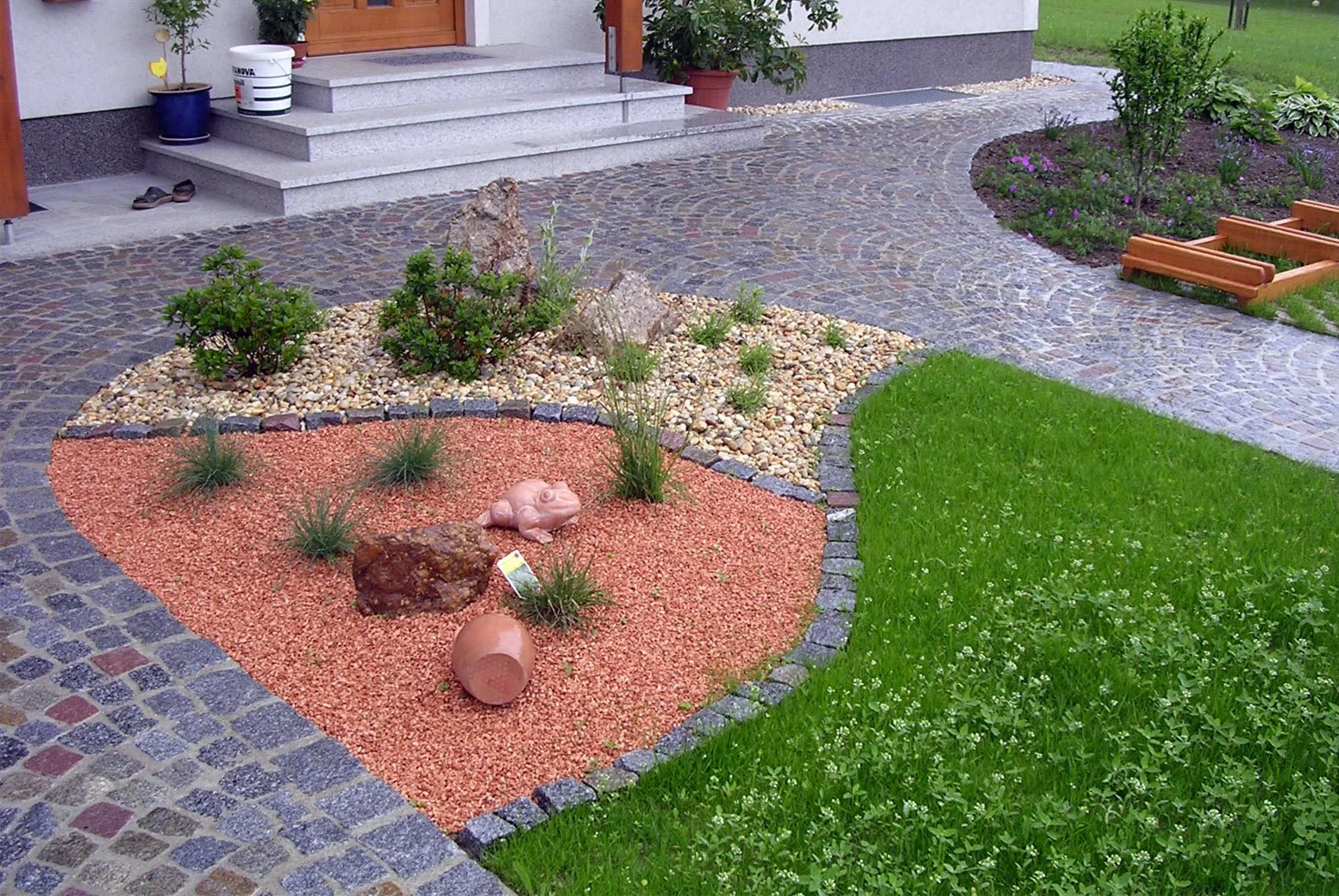 Garten mit verwittertem Terrakottakrug, Steinen, Pflanzen, Kieselsteinfläche, Steinen, Pflastersteinen, Rasen, Blumenbeeten und Eingangsbereich mit Treppe.
