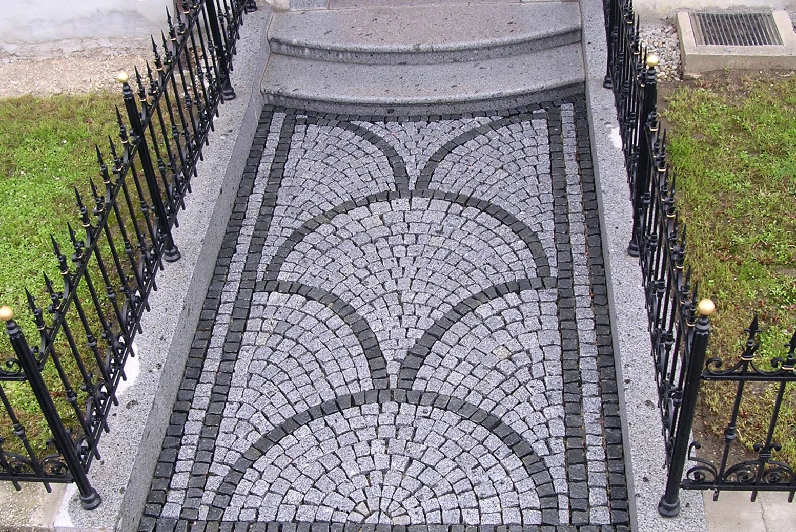 Steinpflaster mit einem Muster aus Halbrundbögen, umgeben von einem Steinrand, eingezäunt von einem schwarzen Metallzaun mit goldfarbenen Knöpfen, vor Treppe.