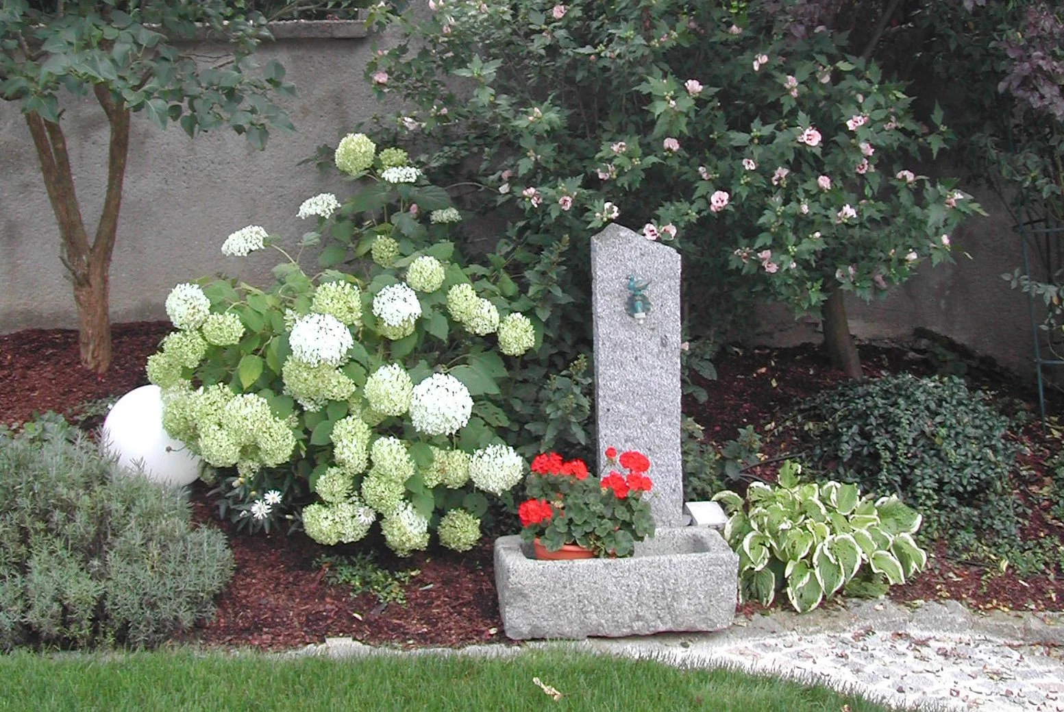 Garten mit verschiedenen Blumen, einem kleinen Springbrunnen, einem Steinpavillon und einem Bewässerungshahn.